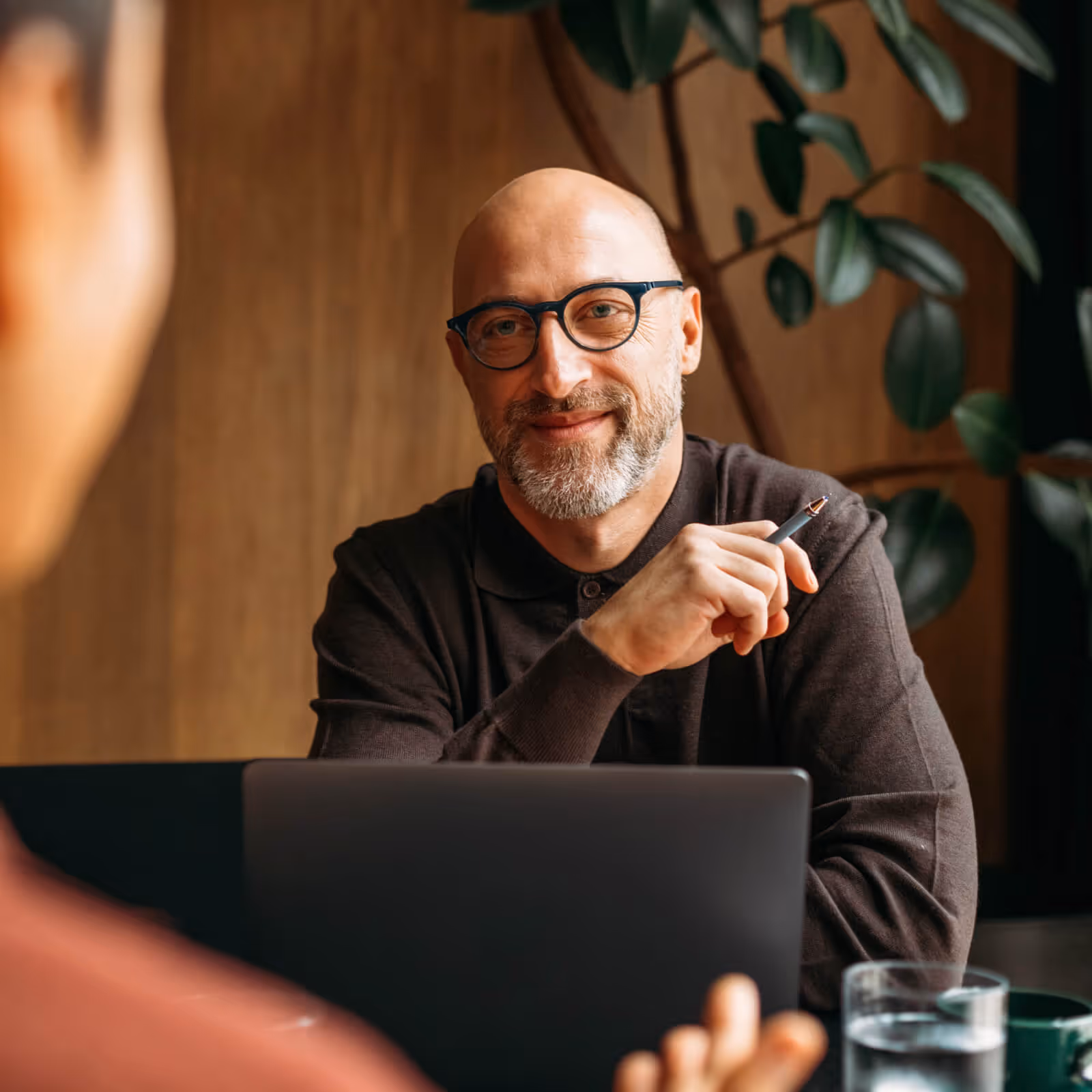 Smiling bald man with glasses holding a pen, sitting at a table with a laptop and talking to another person.