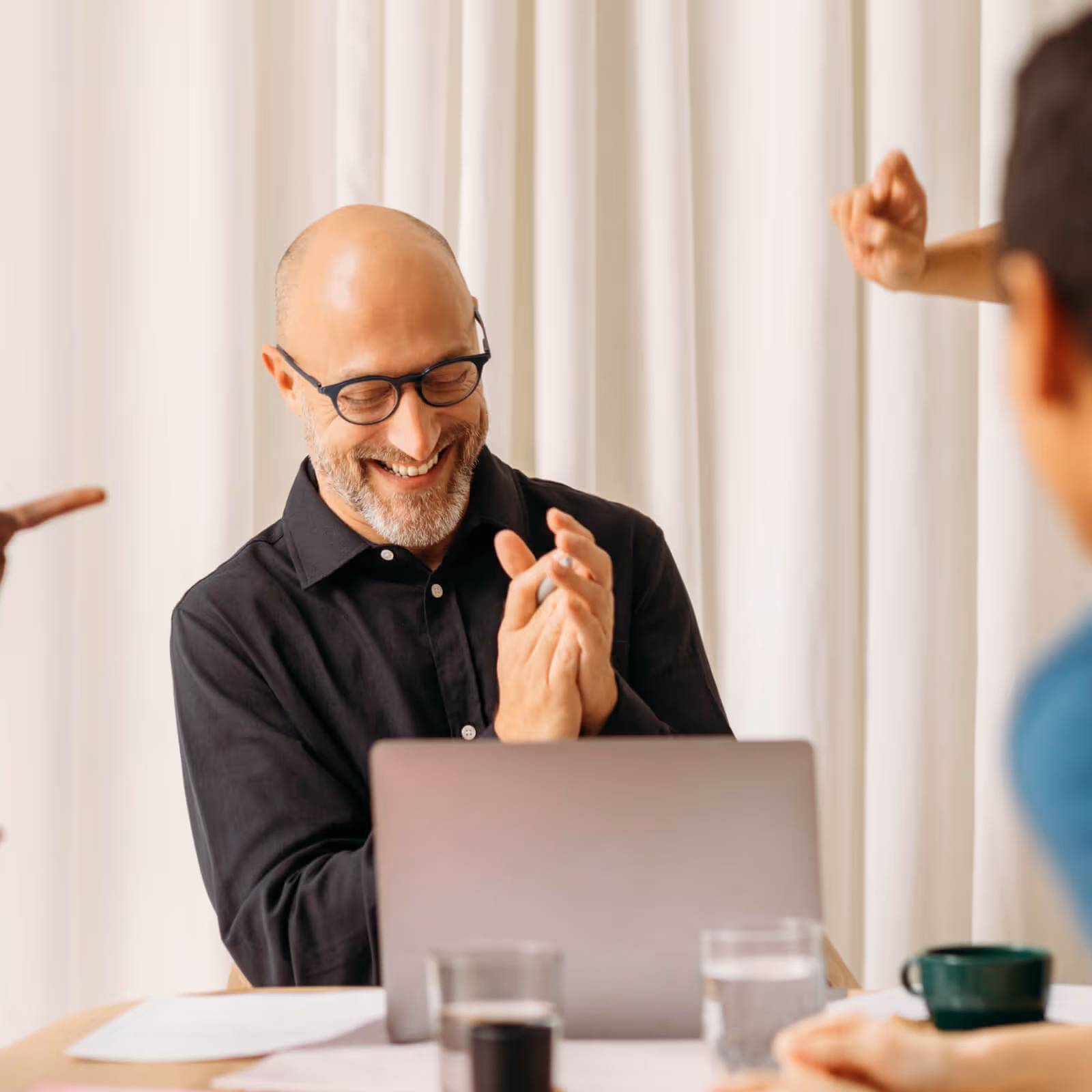 Smiling man with glasses clapping hands in front of a laptop during a meeting.