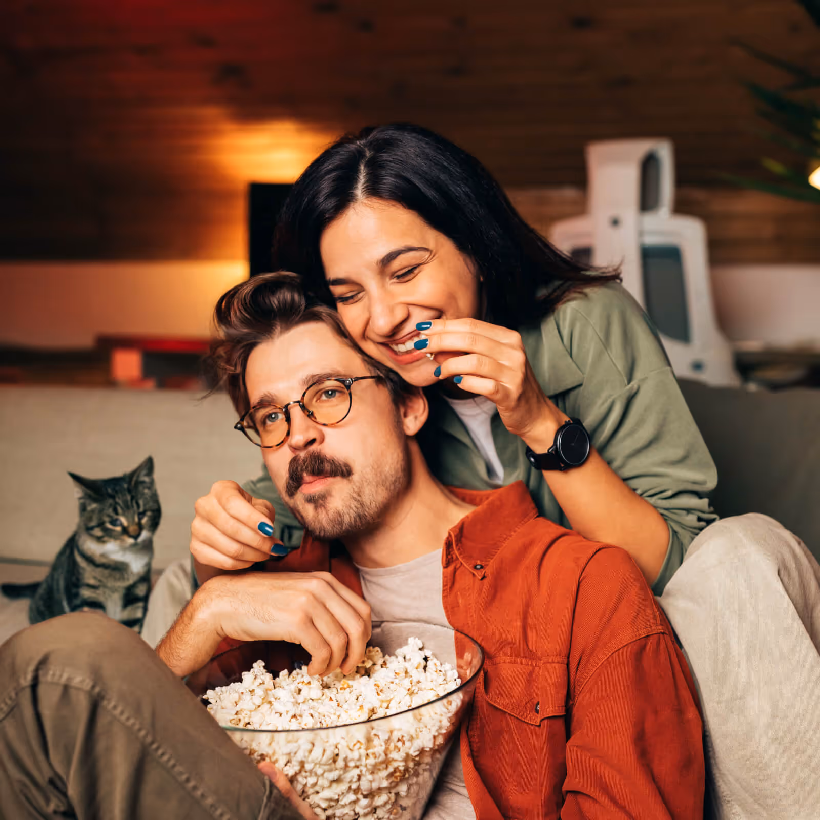 Smiling woman hugs man from behind as they share a bowl of popcorn on a couch with a cat nearby.