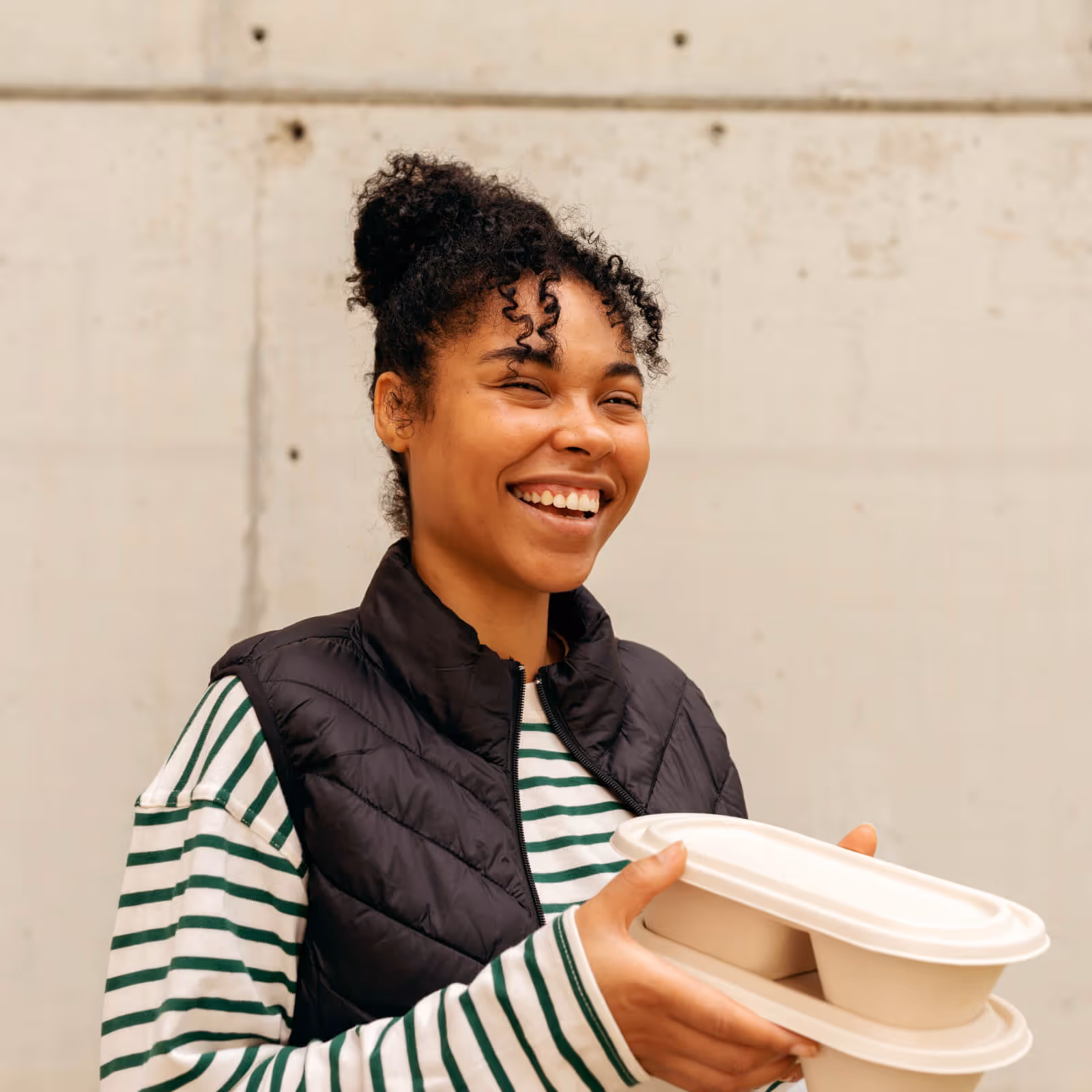 Smiling woman wearing a black vest and striped shirt holding two stacked takeout food containers.