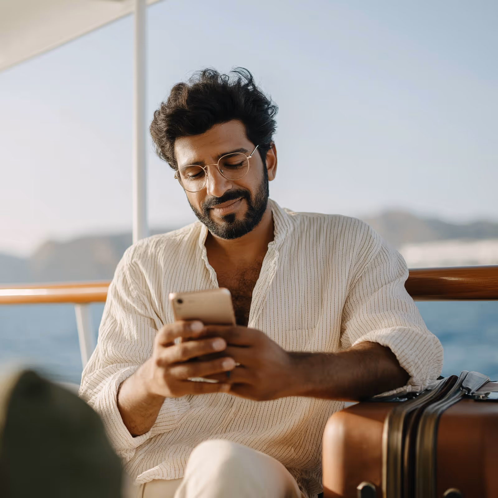 Smiling man wearing glasses and a light shirt sitting by a brown suitcase on a boat, looking at his smartphone.
