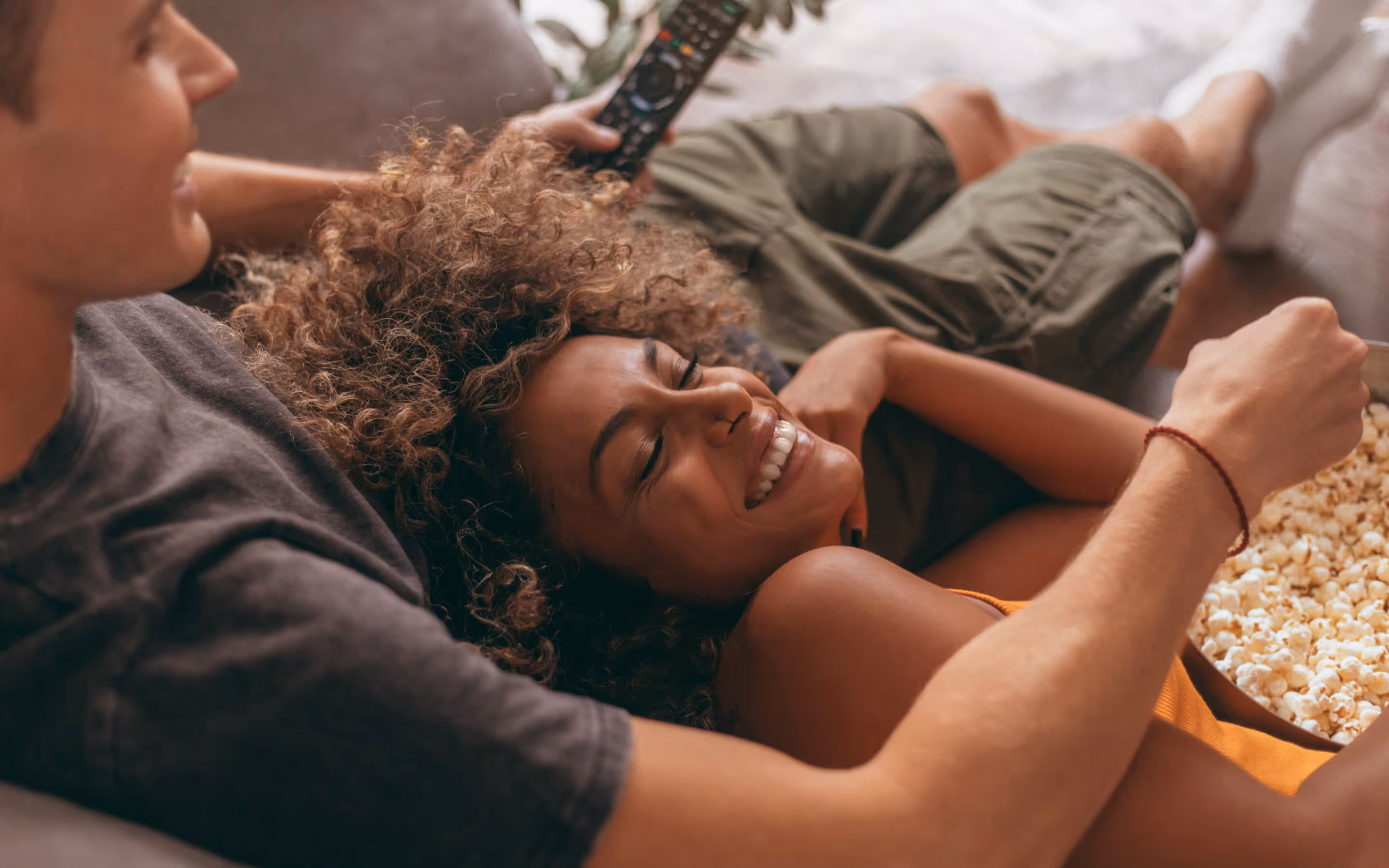 A smiling woman with curly hair lying on a man's lap while he holds a remote and they share a bowl of popcorn.