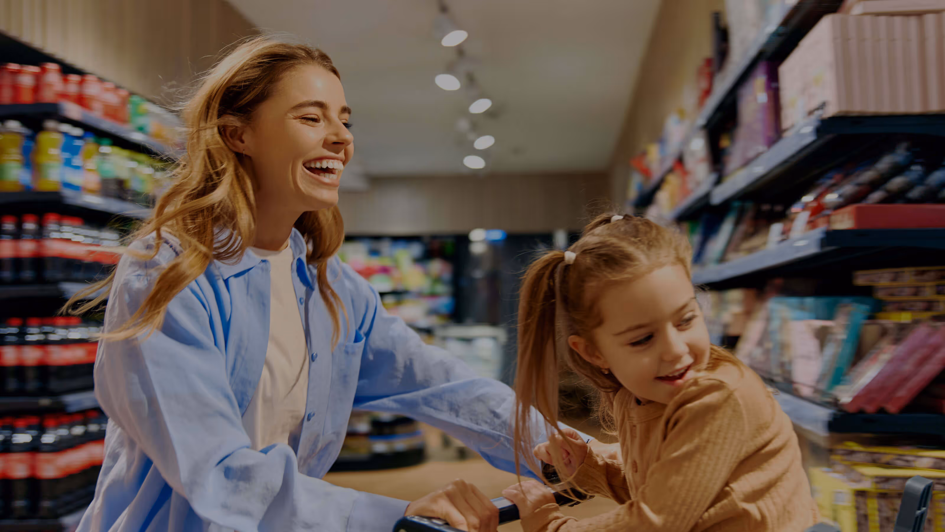 A smiling woman pushes a shopping cart with a cheerful young girl riding inside through a brightly lit grocery store aisle. They both laugh, sharing a joyful moment near shelves stocked with drinks and snacks.