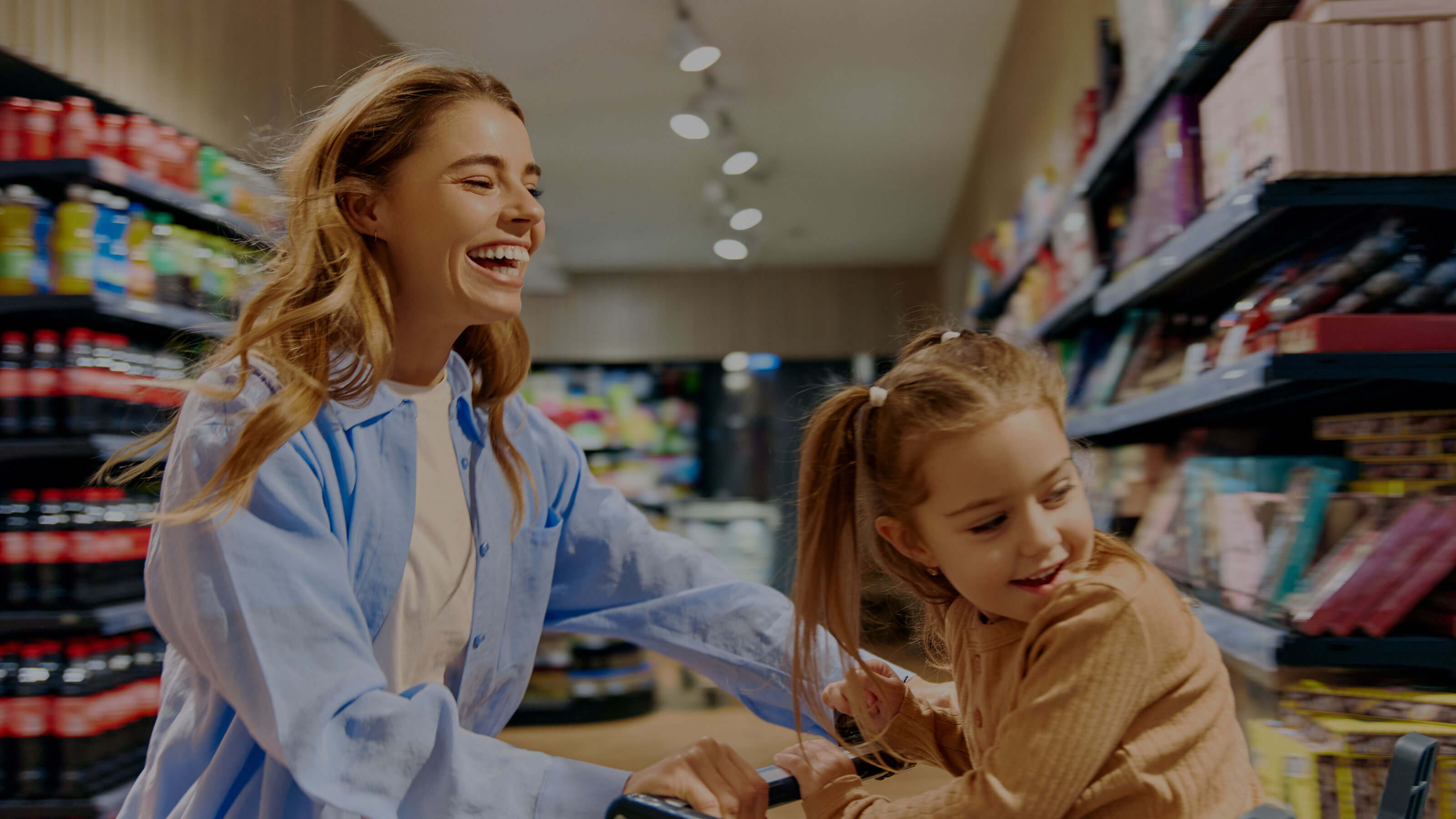 A smiling woman pushes a shopping cart with a cheerful young girl riding inside through a brightly lit grocery store aisle. They both laugh, sharing a joyful moment near shelves stocked with drinks and snacks.