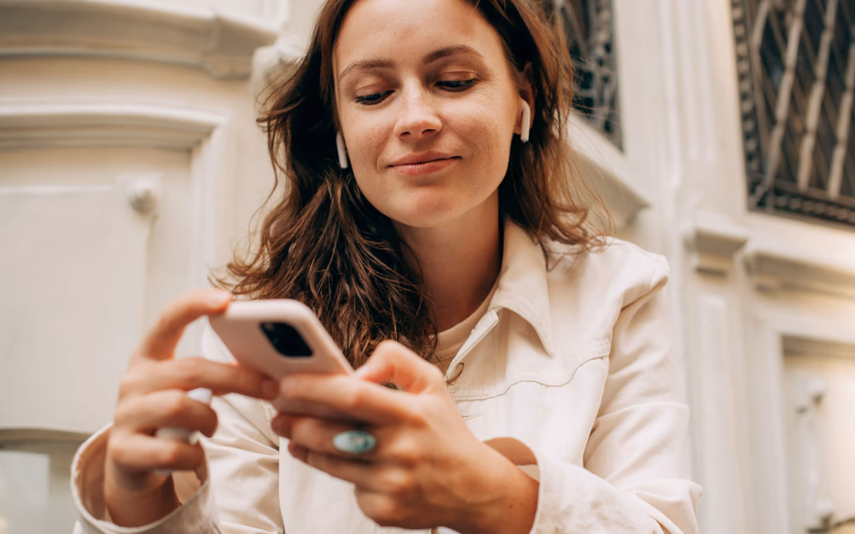 Young woman with earbuds smiling while using a smartphone outdoors.