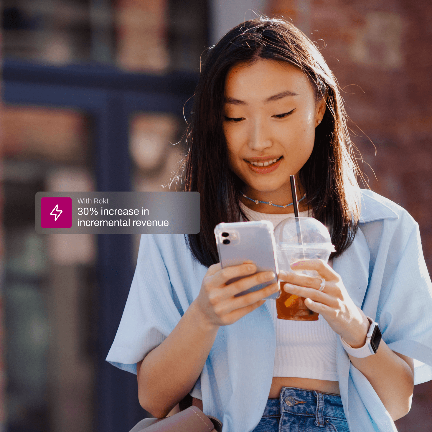 Young woman smiling while looking at her smartphone and holding a plastic cup with iced tea and a straw outdoors.