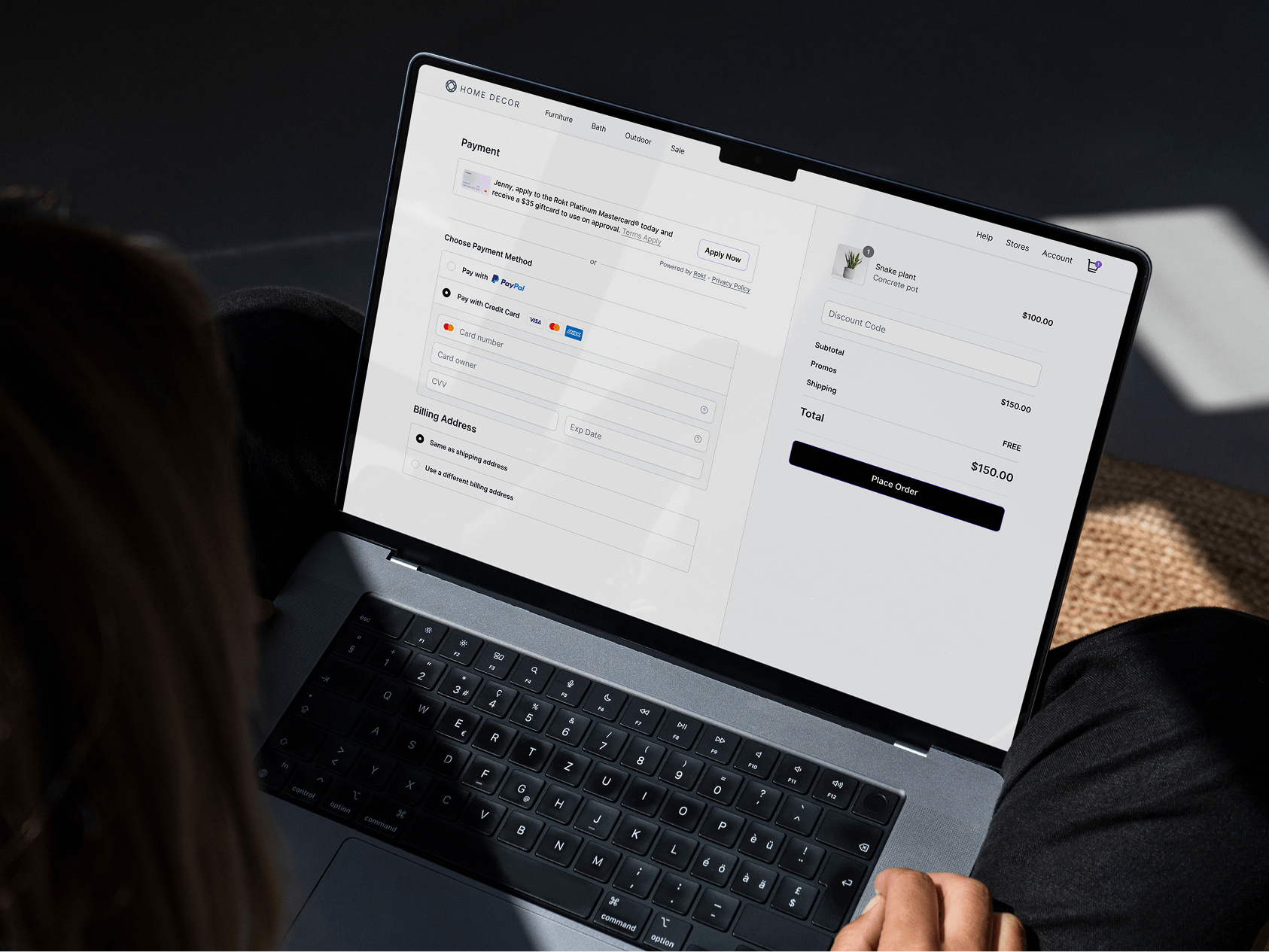 A person uses a laptop to complete an online checkout for a snake plant in a concrete pot on the HOME DECOR website. The screen displays the payment page with options to pay via PayPal or credit card, and a billing address section. On the right, the order summary shows a total of $150 with free shipping, and a prominent “Place Order” button is ready to be clicked. Sunlight casts soft shadows, suggesting a bright indoor setting.