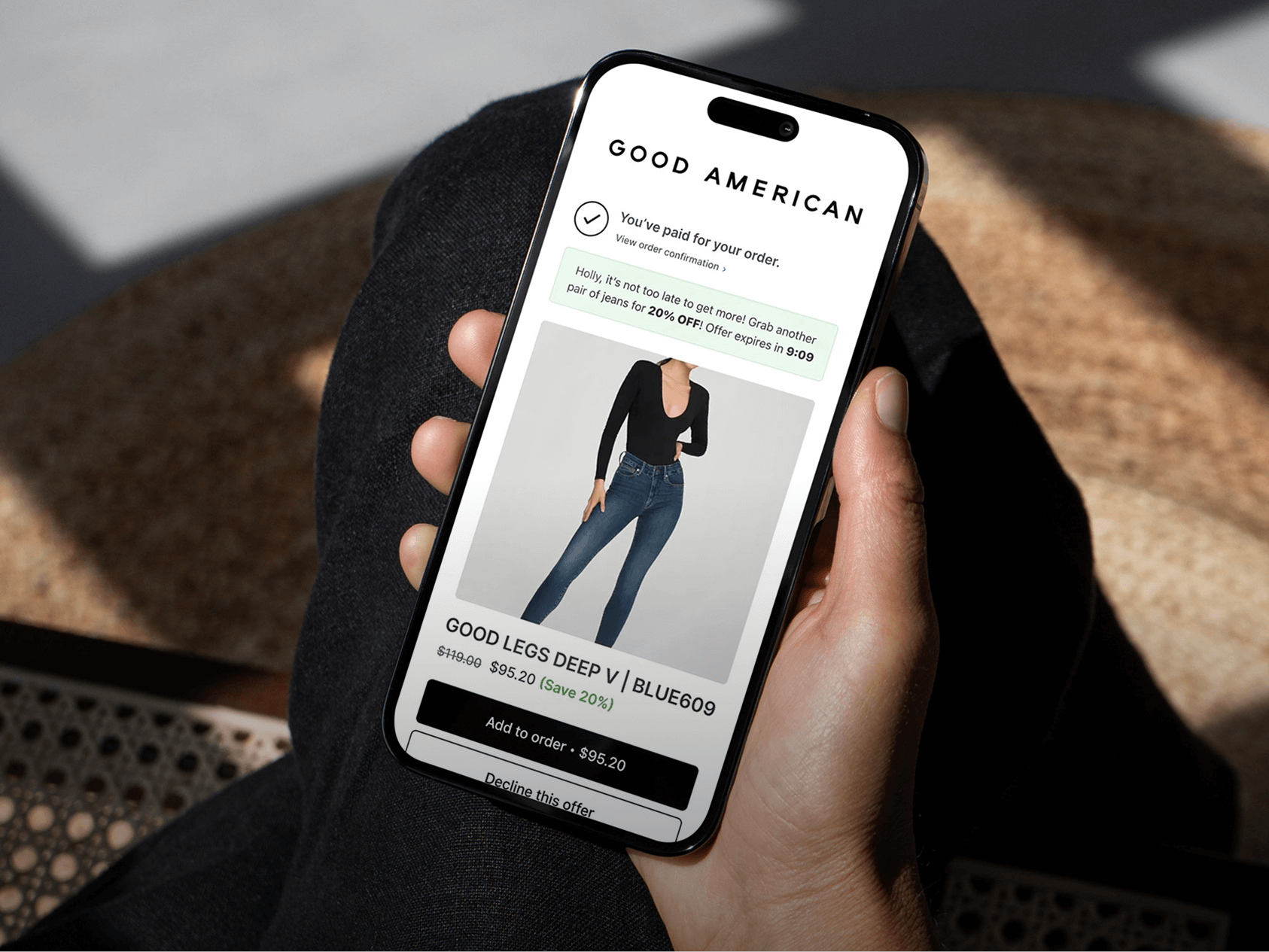 A person holds a smartphone displaying an order confirmation page from the brand GOOD AMERICAN. The screen shows a completed purchase and promotes a limited-time offer to buy an additional pair of “GOOD LEGS DEEP V | BLUE609” jeans at 20% off. The jeans are modeled by a woman wearing a black long-sleeve top and high-waisted denim. The interface includes buttons to “Add to order” or “Decline this offer,” and the scene is brightly lit with natural sunlight.