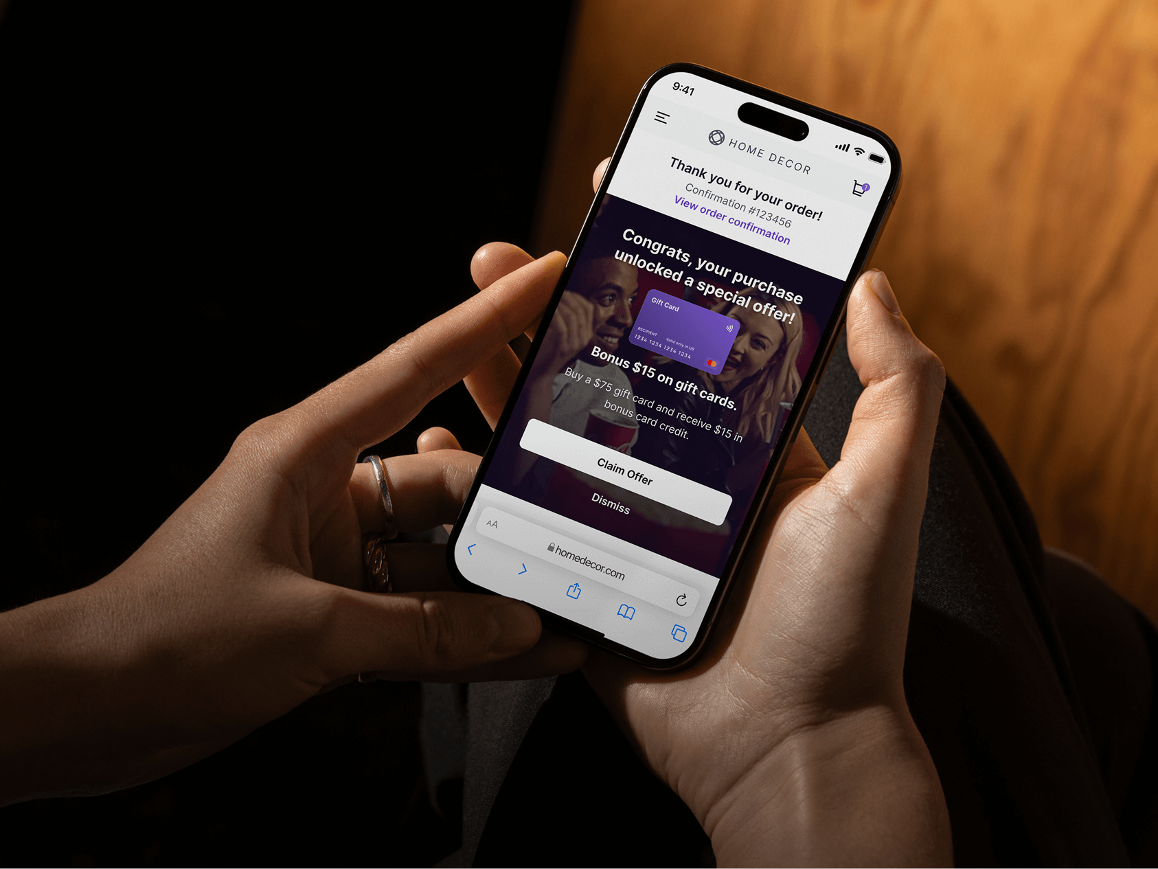 A person holds a smartphone displaying a post-purchase confirmation screen from the HOME DECOR website. The message thanks the user for their order and promotes a bonus offer: “Congrats, your purchase unlocked a special offer!” offering $15 in bonus credit when buying a $75 gift card. Below the message are “Claim Offer” and “Dismiss” buttons. The phone is held with both hands in a dimly lit, warm-toned environment, and the screen is brightly illuminated.