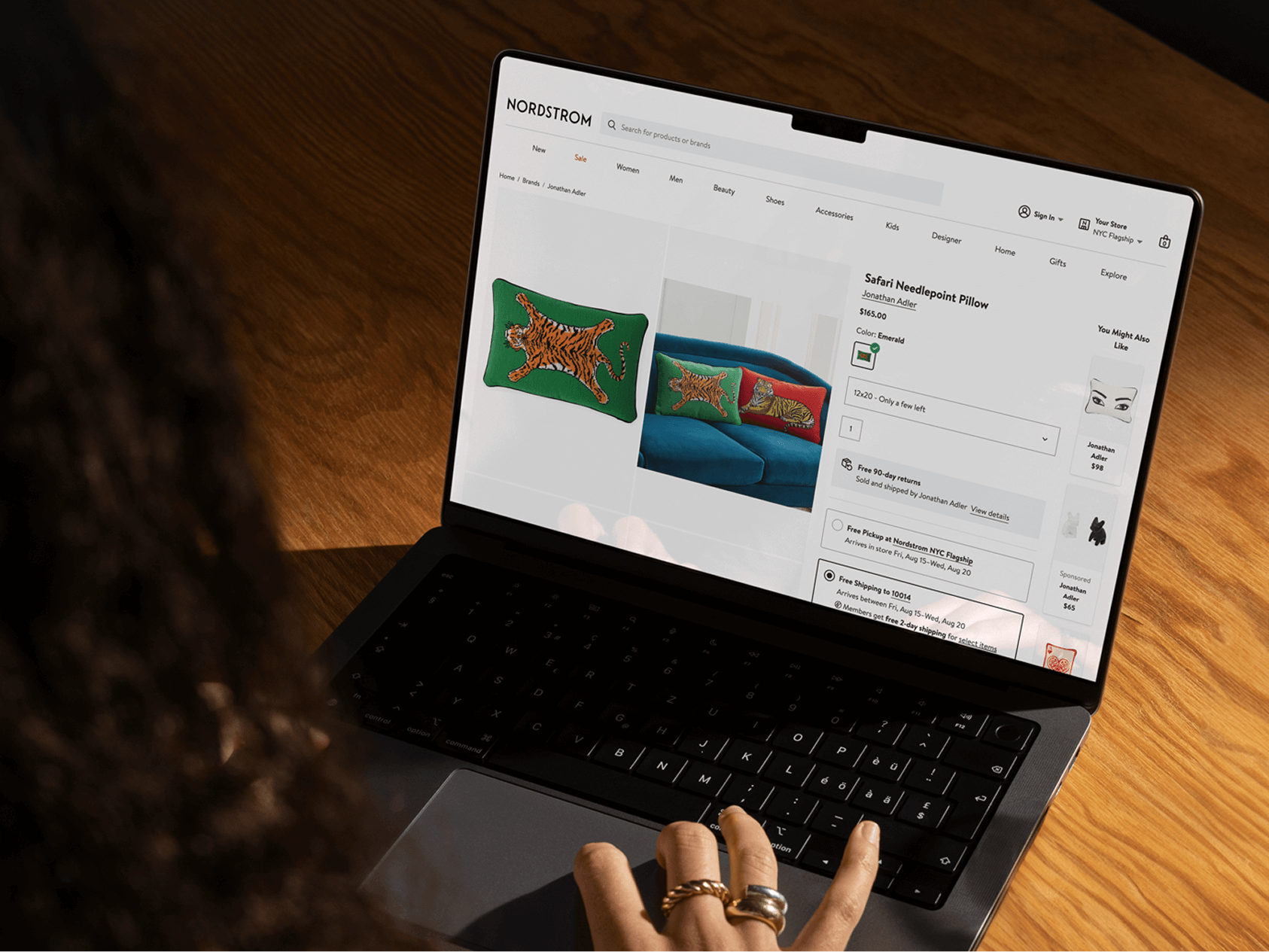 A person browses the Nordstrom website on a laptop, viewing a product page for a “Safari Needlepoint Pillow” by Jonathan Adler. The pillow features a colorful embroidered tiger on a green background and is shown both as a standalone item and on a blue couch in a lifestyle image. The selected color is "Emerald," and delivery and pickup options are visible. The scene is warmly lit, with the user's hand on the keyboard and wood grain visible on the tabletop.