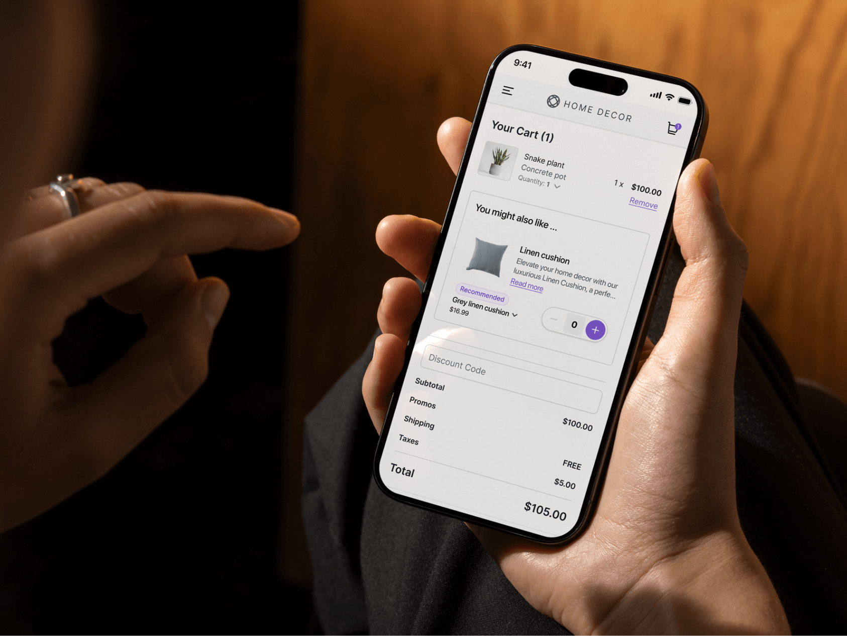 A person holds a smartphone displaying the shopping cart page from the HOME DECOR website. The cart contains one item—a snake plant in a concrete pot priced at $100. Below, a suggested item appears: a grey linen cushion with an option to add it to the cart for $16.99. The order summary at the bottom shows free shipping, $5 in taxes, and a total of $105. A second hand points toward the screen, and the setting is warmly lit with a wooden surface in the background.