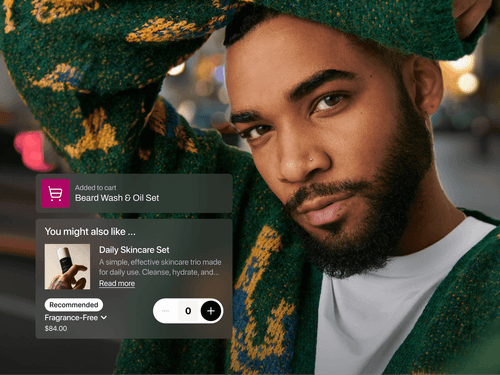 A man with a neatly groomed beard and nose piercing, wearing a green sweater with yellow patterns, poses confidently in an urban night setting. Overlaid is a shopping notification showing he added a “Beard Wash & Oil Set” to his cart. A recommendation for a $84 Daily Skincare Set appears below, with a quantity selector and a dropdown for fragrance options.