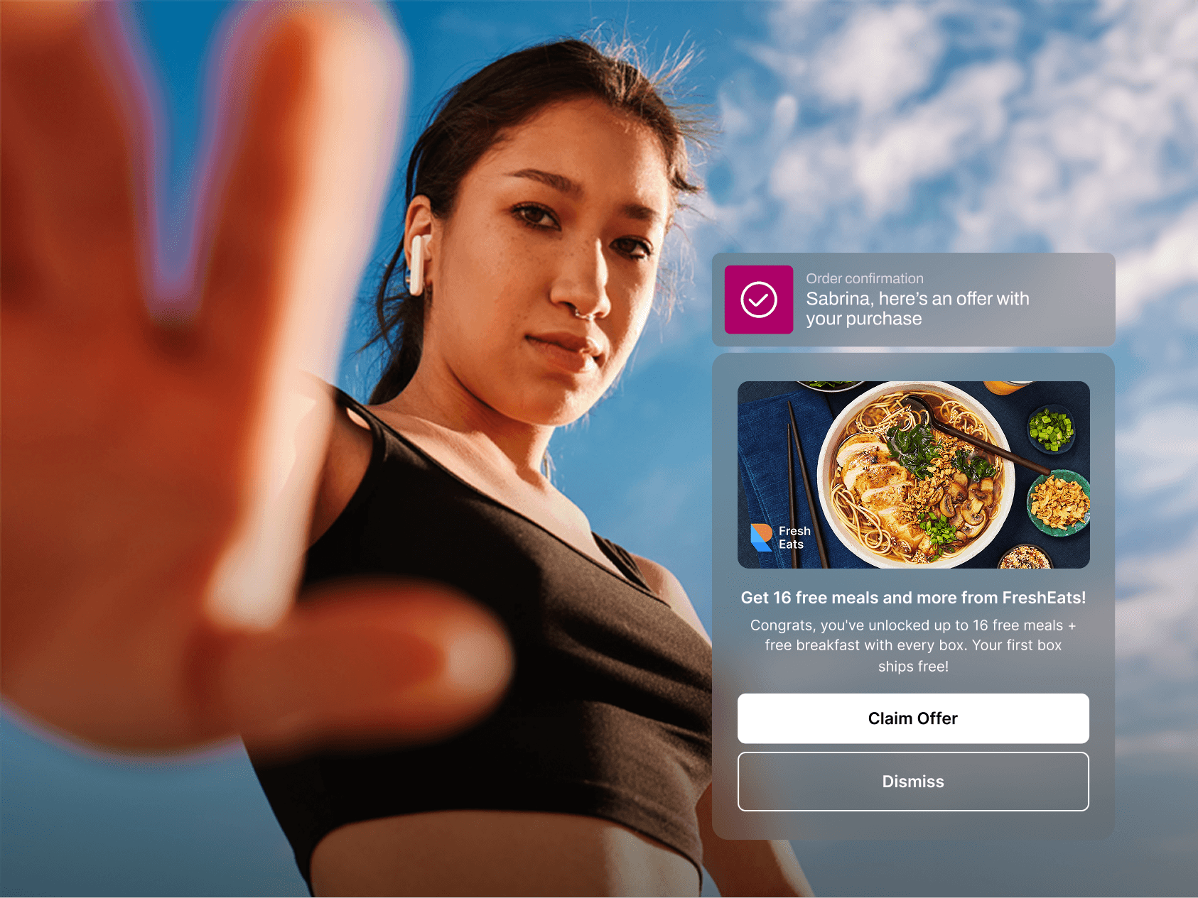 A young woman in athletic wear reaches toward the camera with one hand, creating a blurred foreground effect. She wears wireless earbuds and looks confidently into the lens against a bright blue sky. Overlaid is an order confirmation for “Sabrina,” promoting a FreshEats offer: up to 16 free meals and free breakfast with each box. A food image shows a bowl of ramen with chopsticks and side dishes. Buttons below read “Claim Offer” and “Dismiss.”