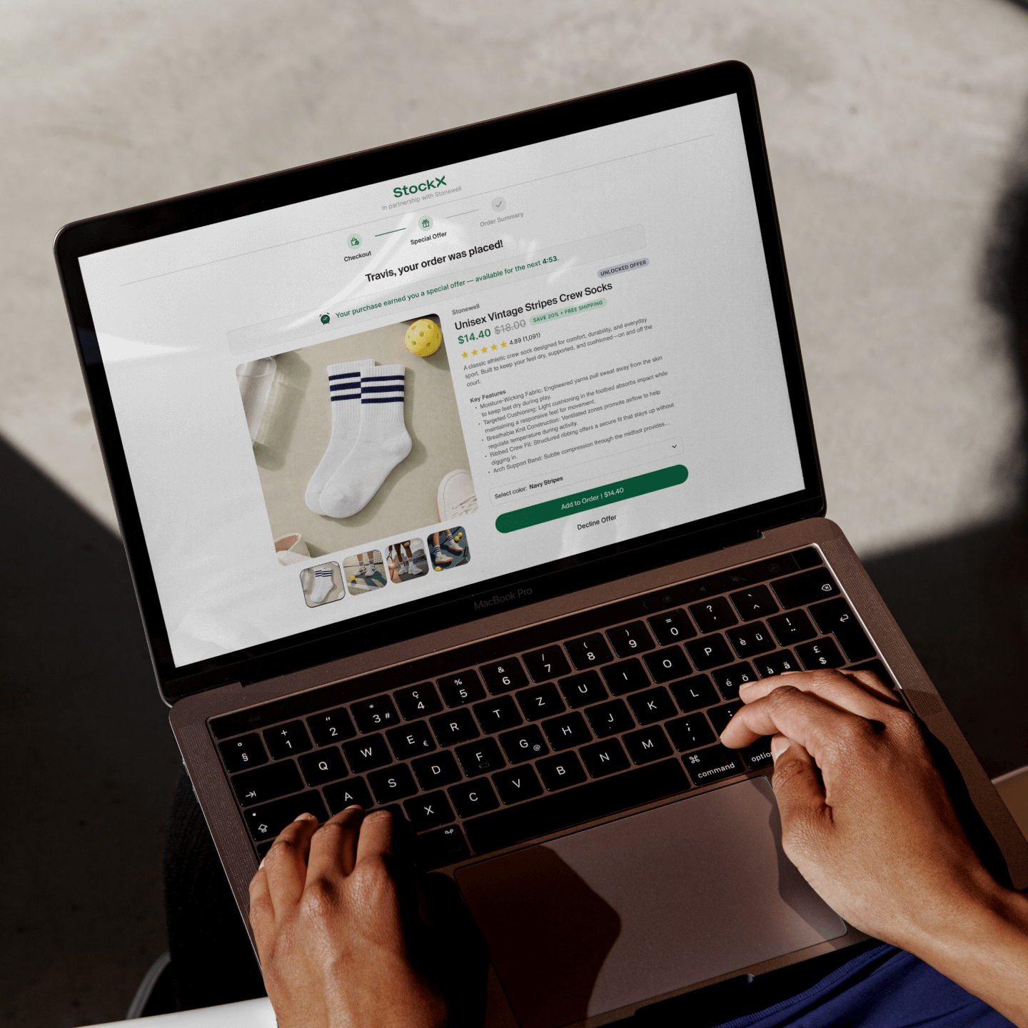 A person using a MacBook Pro places an order on the StockX website for a pair of unisex vintage striped crew socks. The screen displays a confirmation message for the purchase and an upsell offer for additional socks. The featured product is a pair of white crew socks with three navy blue stripes near the cuff, shown alongside lifestyle photos. The user's hands are on the keyboard, and natural sunlight casts shadows on the laptop and nearby surface.