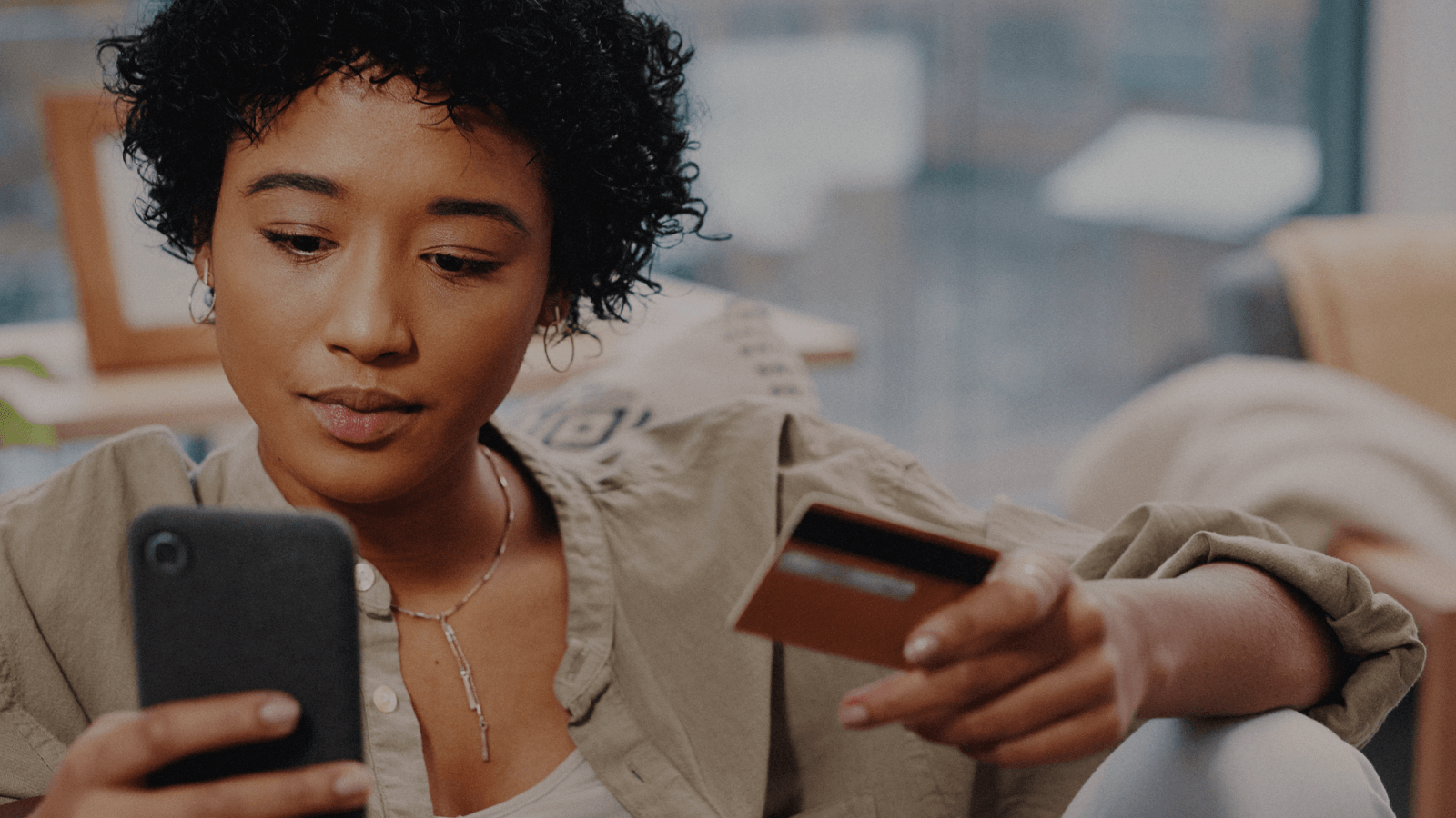 A young woman sits indoors, focused on her smartphone while holding a credit card, suggesting an online purchase or mobile banking. Soft natural light and a casual home setting emphasize everyday digital payments and financial management.