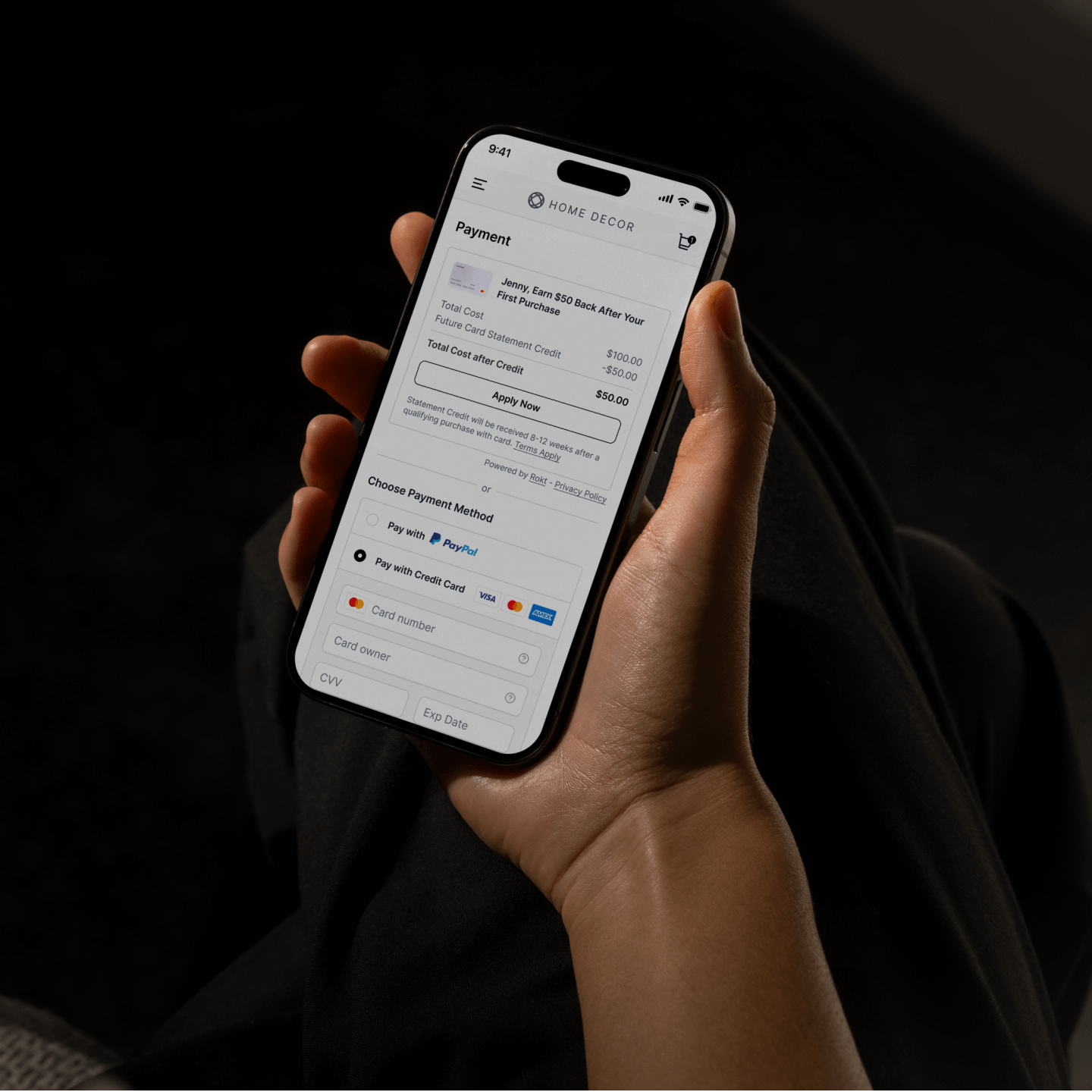 A hand holds a smartphone displaying a mobile checkout screen for a home décor purchase, showing payment options, credit card details, and a promotional credit. The dark background and focused framing emphasize secure online shopping and digital payments.