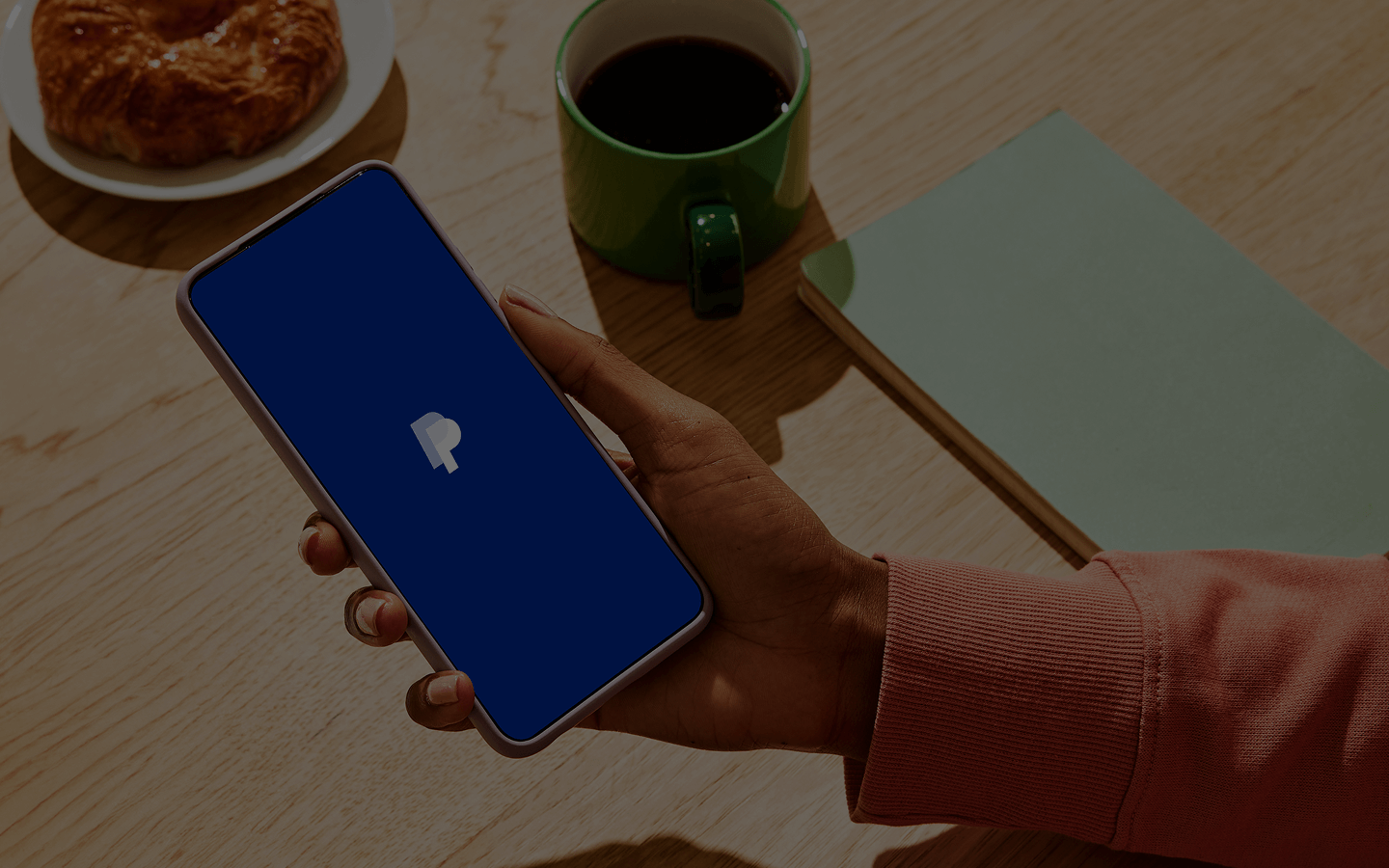 A person in a rust-colored sweater holds a smartphone showing the PayPal logo on a blue screen, suggesting a digital payment or finance app. The phone is held above a light wooden table set with a green mug of black coffee, a croissant on a white plate, and a closed sage-green notebook, creating a casual morning workspace scene.
