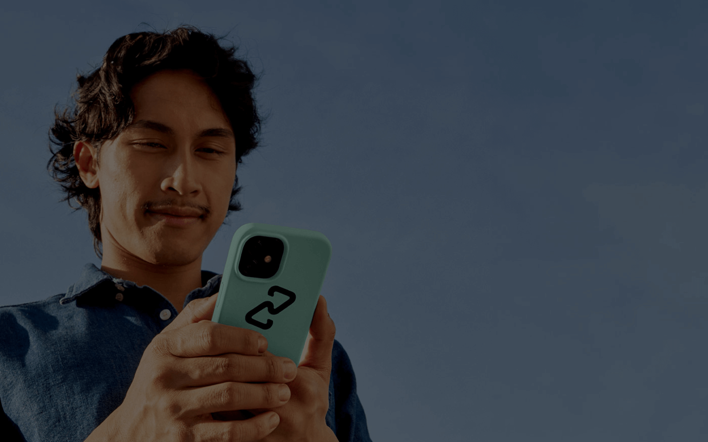 A young man with medium-length dark hair and a faint mustache stands outside beneath a cloudless blue sky. He wears a dark blue collared shirt and holds a smartphone in both hands, looking down at the screen with a calm, slight smile. The phone has a mint-green case with a black graphic design on the back. The image is shot from a low angle, making the sky fill the background and giving the scene a bright, open, daytime feel.