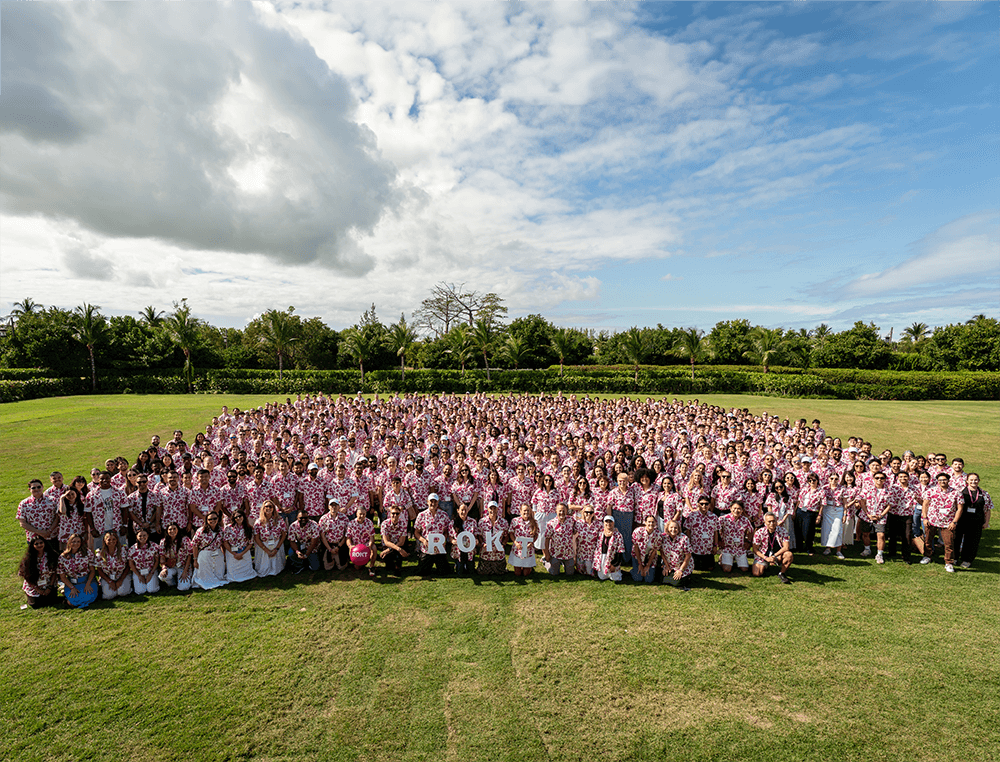 Large group of people wearing matching white and red floral shirts assembled outdoors on green grass under a partly cloudy blue sky.