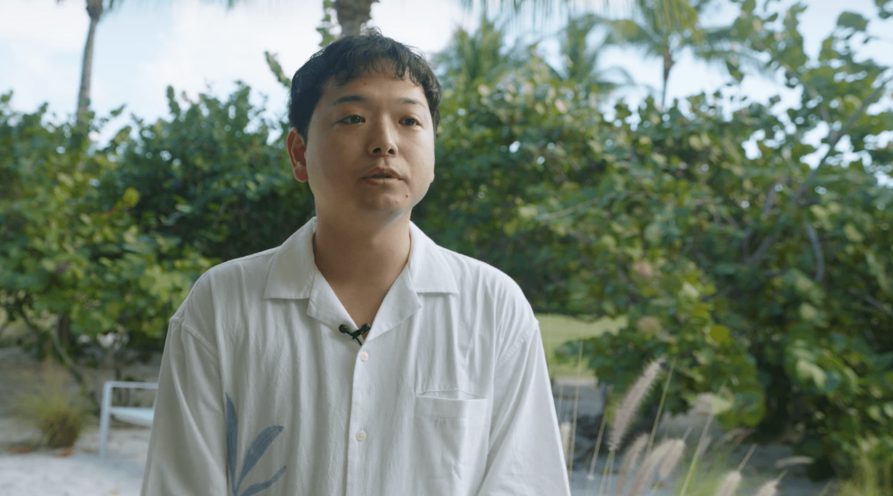 Man in a white shirt with a microphone clipped on, standing outdoors with green foliage and trees in the background.