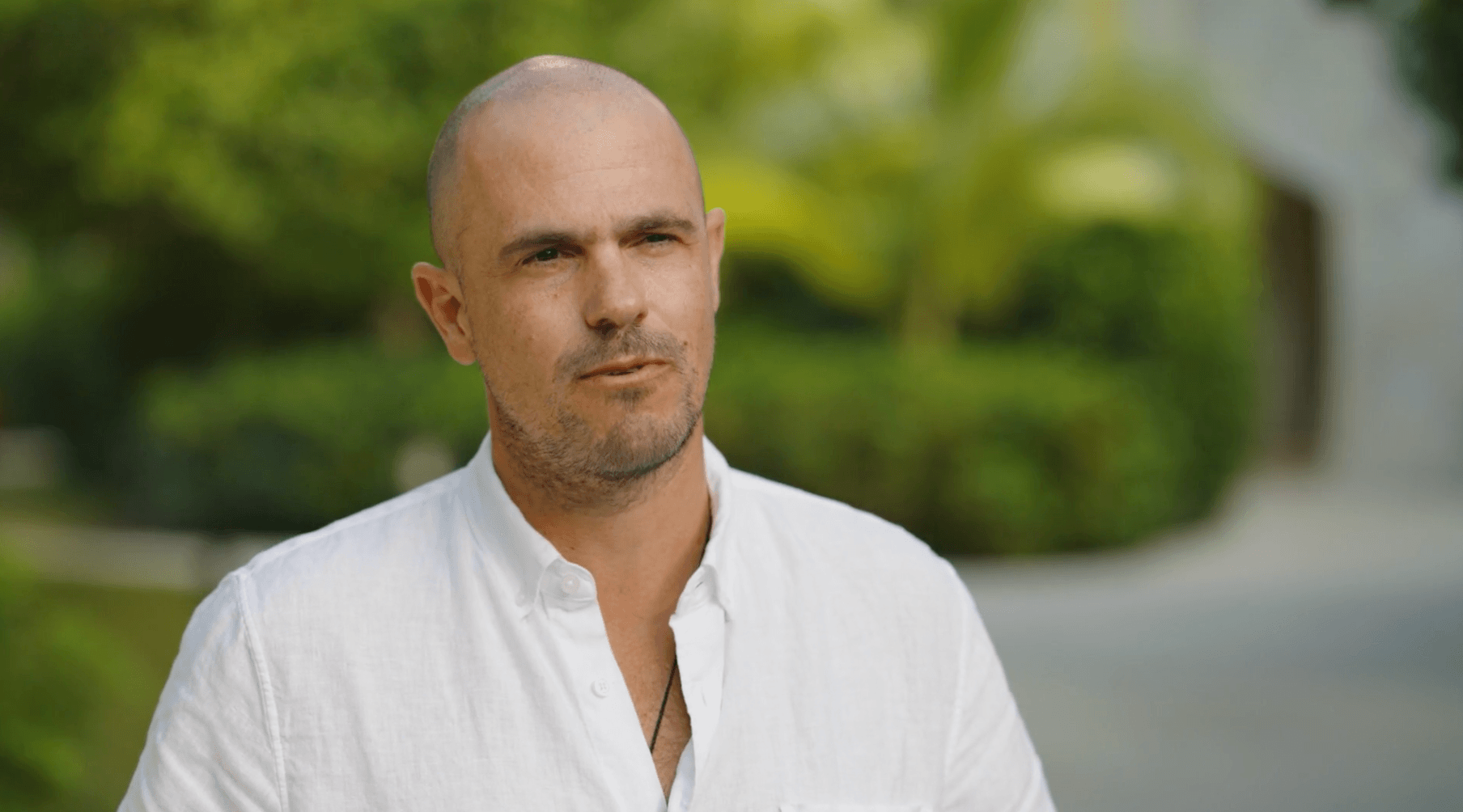 Man with a shaved head and light stubble wearing a white buttoned shirt outdoors with green foliage in the background.