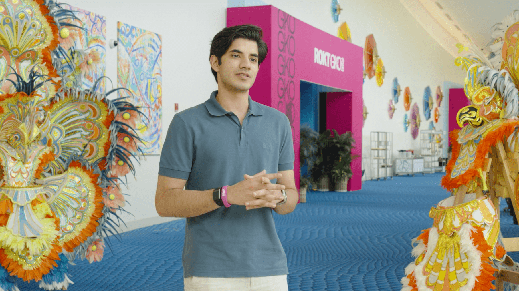 Young man in a blue polo shirt standing in a corridor decorated with colorful feathered costumes and artistic wall decorations.