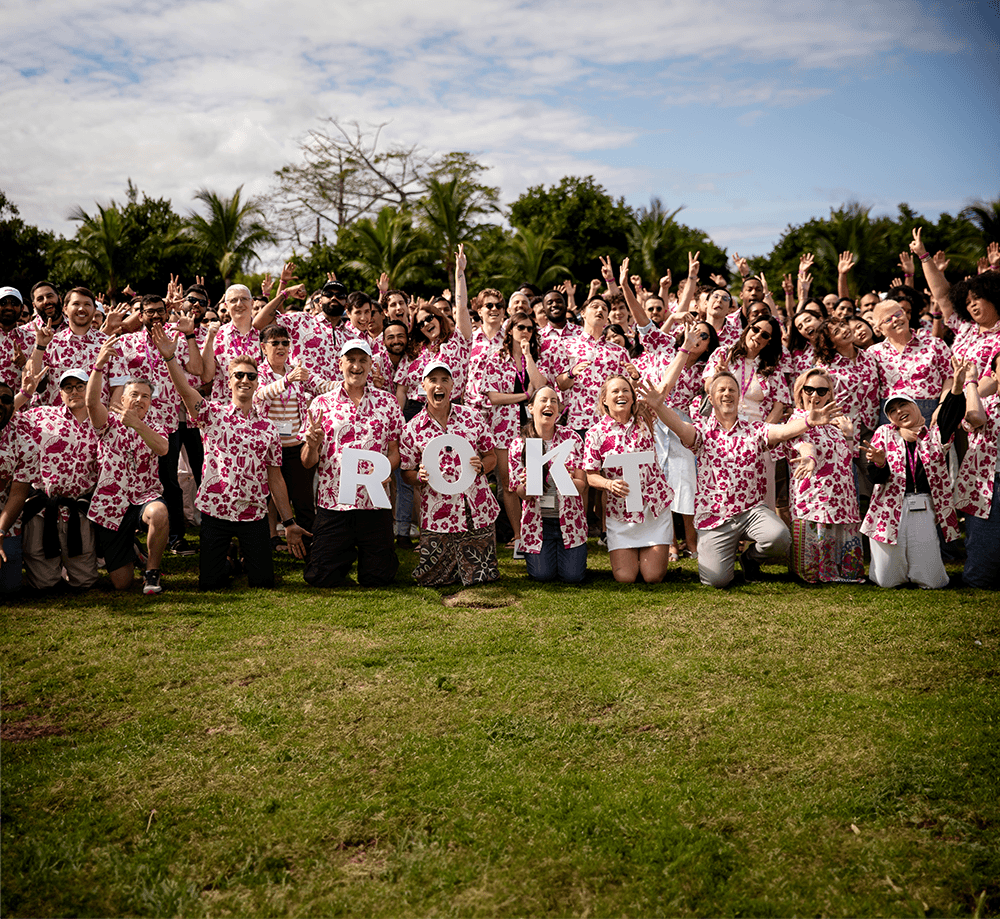 Large group of people in matching pink floral shirts posing outdoors on grass with some holding letters spelling ROKT.