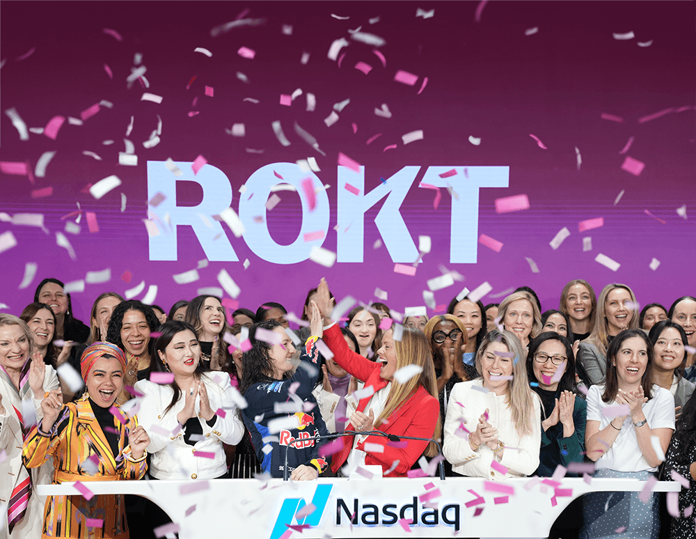 Group of diverse, smiling women celebrating with confetti in front of a Nasdaq podium and a large ROKT logo on a purple background.
