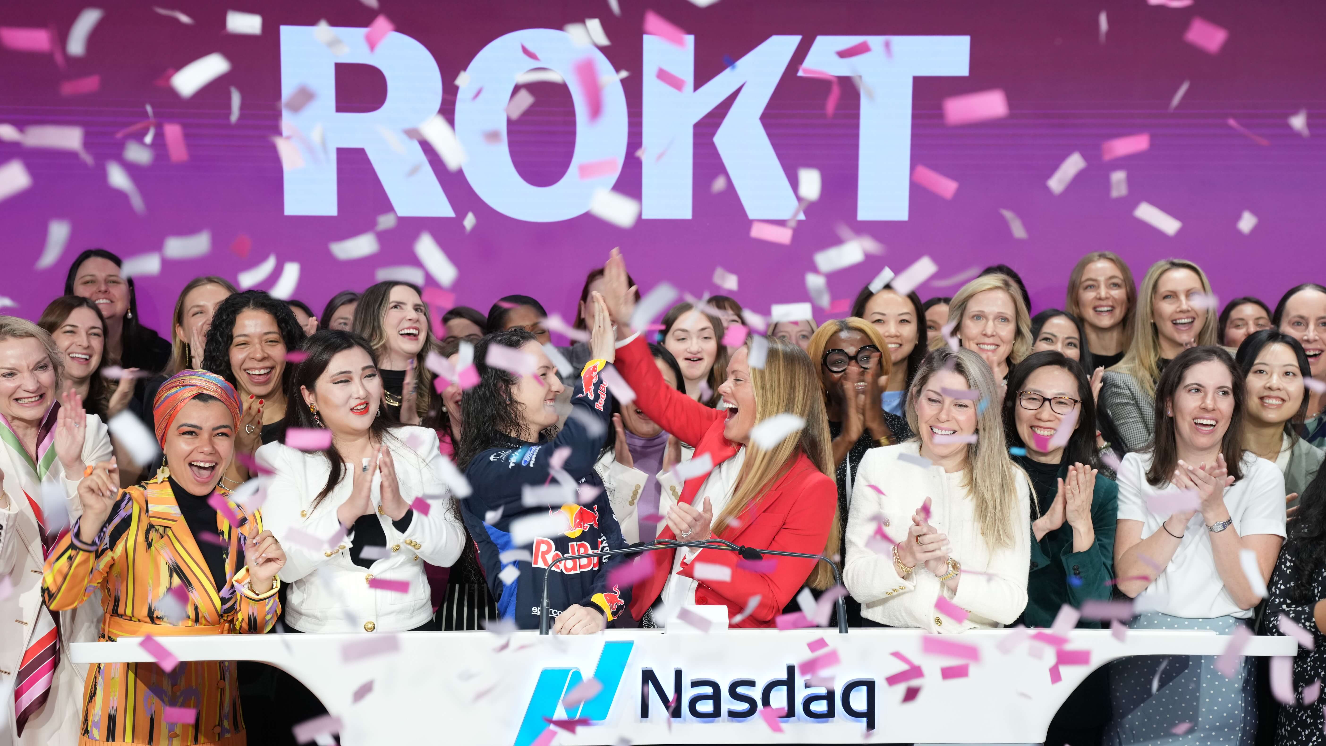 A diverse group of women celebrating with pink and white confetti at a Nasdaq event with a purple 'ROKT' sign in the background.
