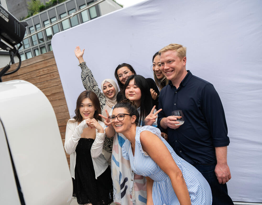 Group of six smiling young adults posing and making playful gestures for a photo against a white backdrop at an outdoor event.