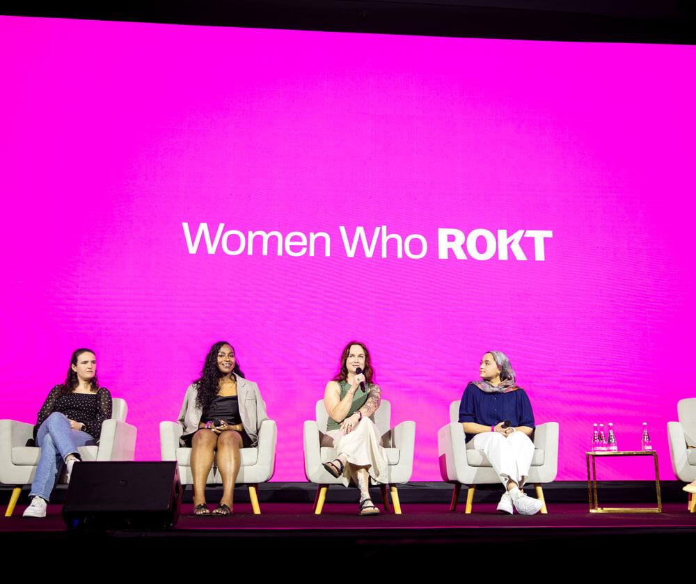Four women seated on stage chairs with a bright pink backdrop displaying the text 'Women Who ROKT' during a panel discussion.