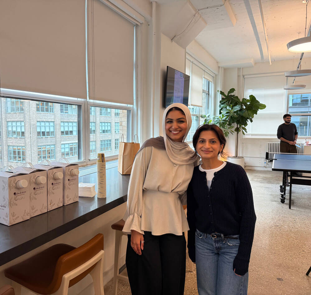 Two women smiling and posing together in a bright office space with large windows and a ping pong table in the background.