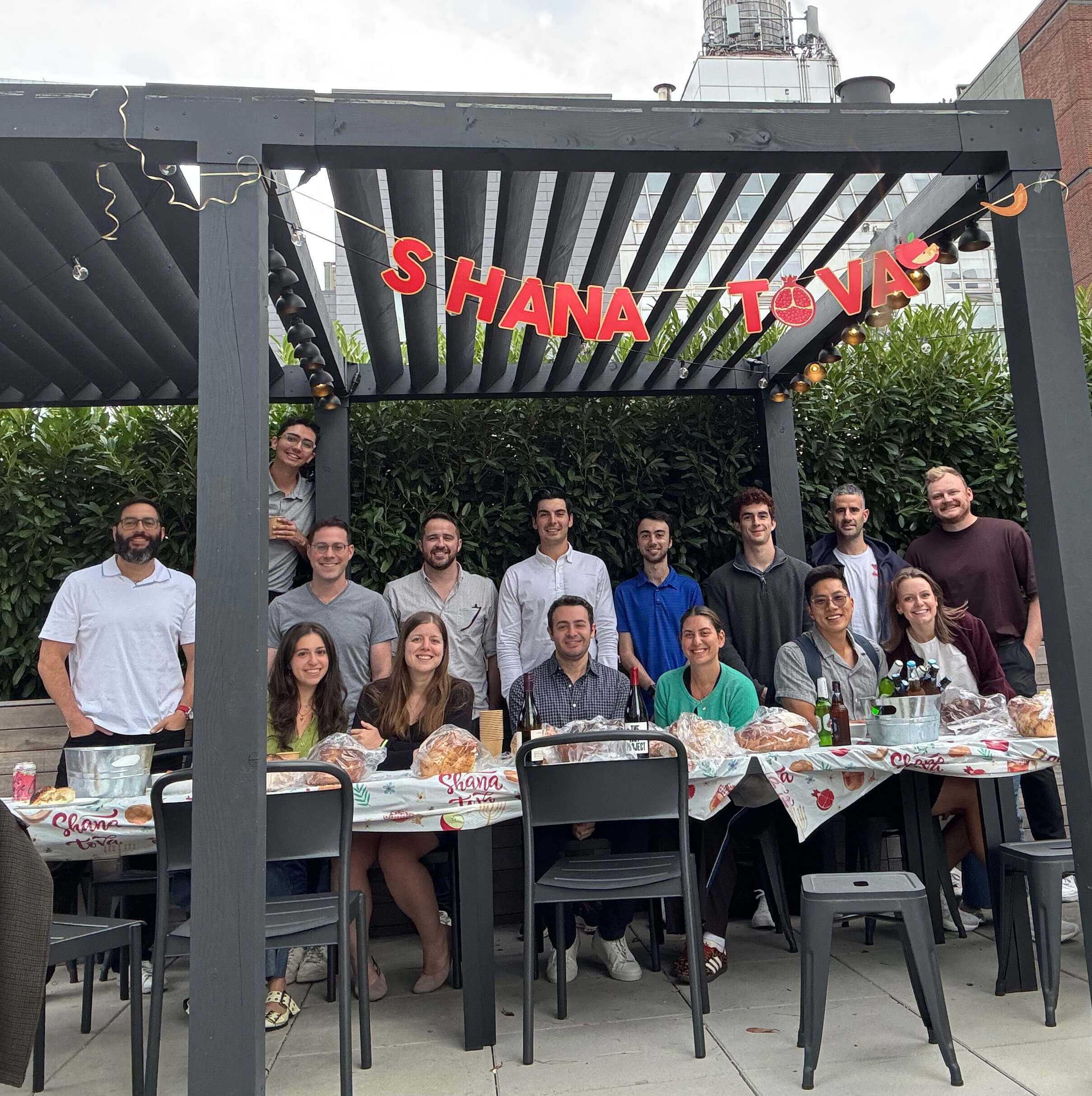 Group of people smiling under a pergola with 'Shana Tova' banner, sitting and standing around a table with challah bread and drinks.