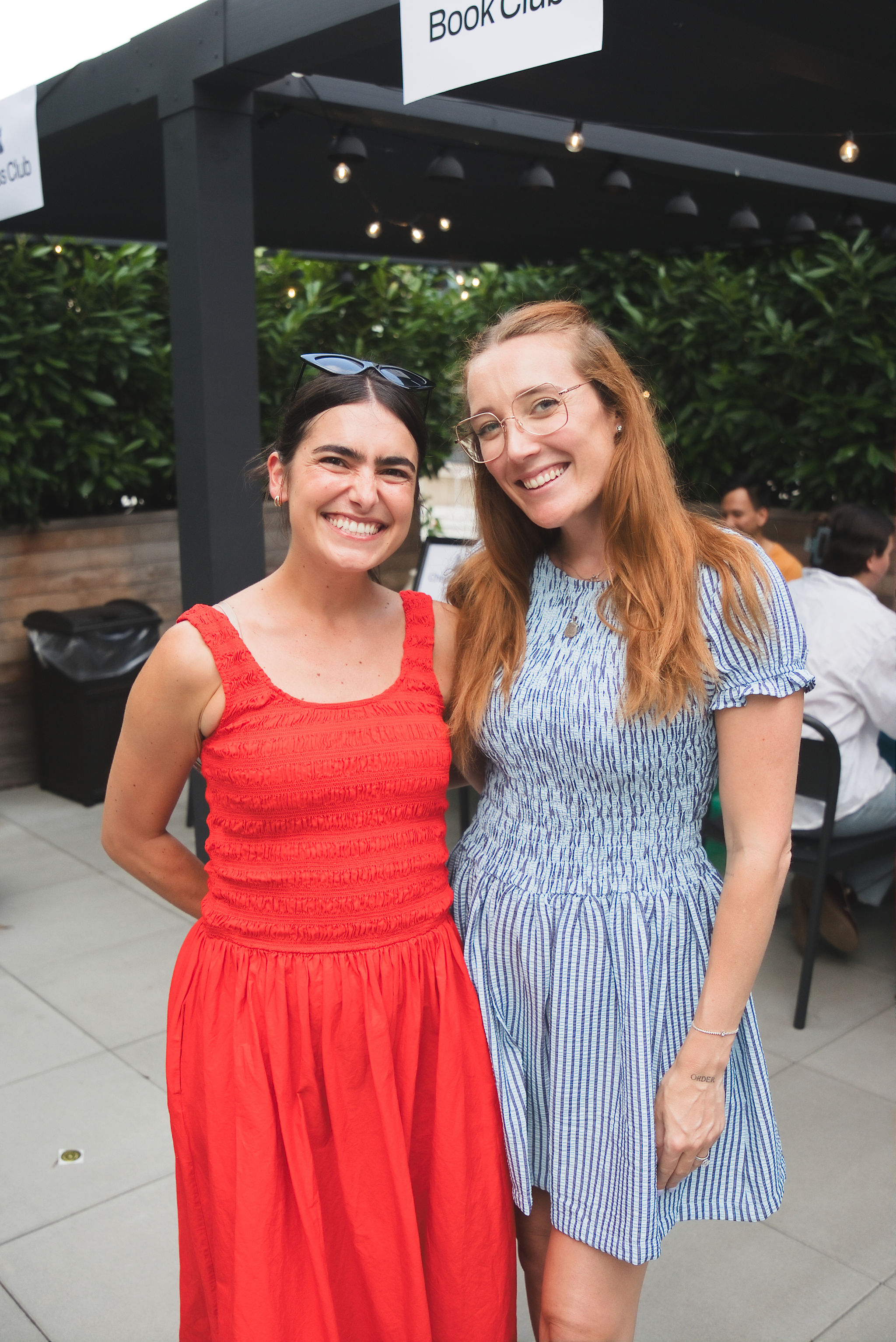 Two smiling women standing outdoors; one in a red dress, the other in a blue and white striped dress.