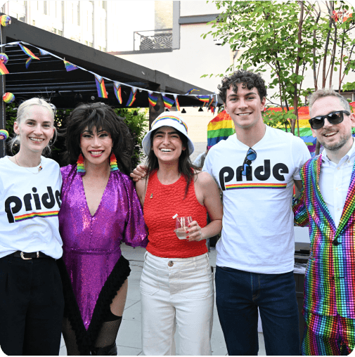 Five people smiling at a Pride event, with colorful rainbow outfits and decorations.