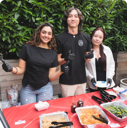 Three people smiling behind a table with food trays, wearing gloves, outdoors with green plants.