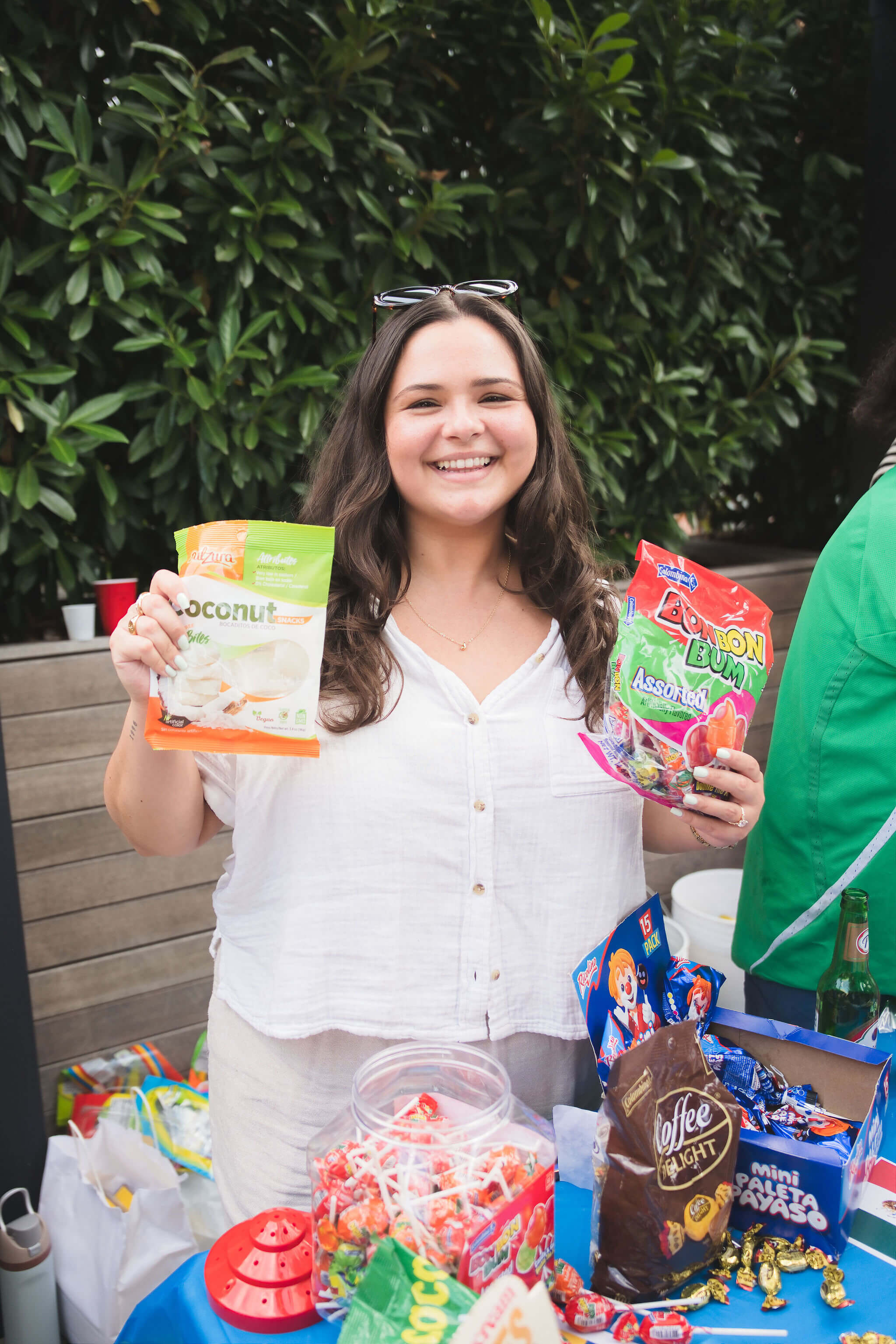 Smiling woman holding two bags of snacks, standing behind a table with assorted candy and treats.