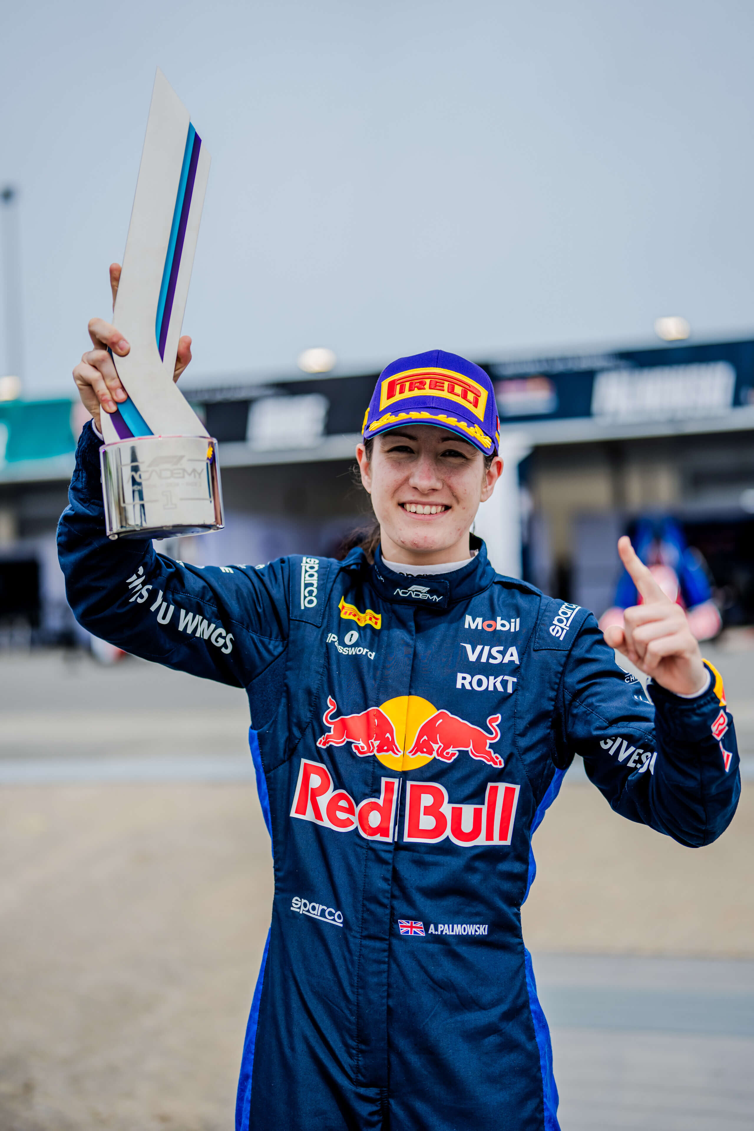 Race car driver in a Red Bull suit holding a trophy and showing a number one gesture.