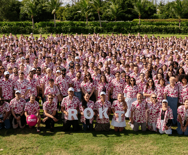 Large group of people outdoors on grass, all wearing matching white shirts with red floral patterns, some holding letters spelling ROKT and a heart-shaped sign with ROKT logo, palm trees and greenery in background.