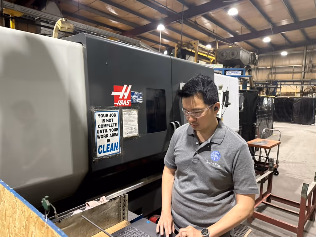 Yen wearing safety glasses and gray SensFlo polo shirt working on a laptop next to a large Haas industrial machine in a factory setting.