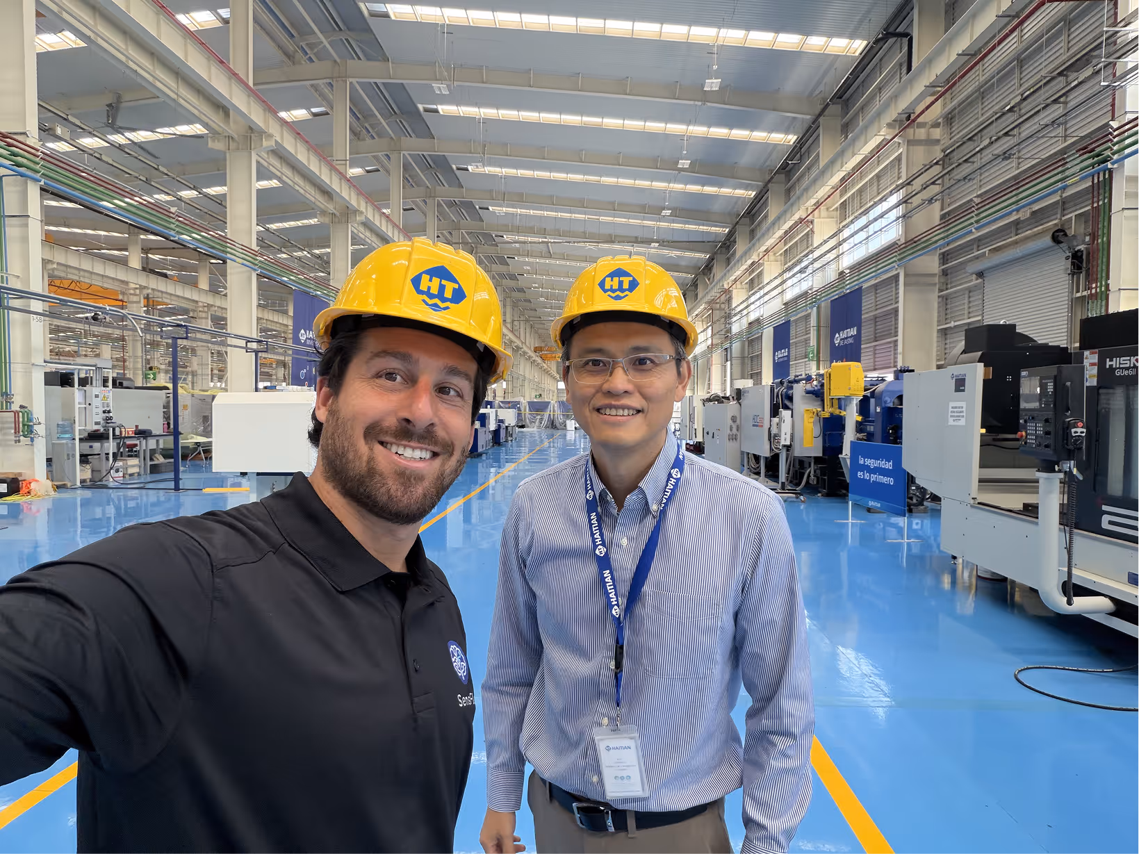 Two men wearing yellow HT hard hats smiling and taking a selfie inside a spacious industrial factory with machinery.