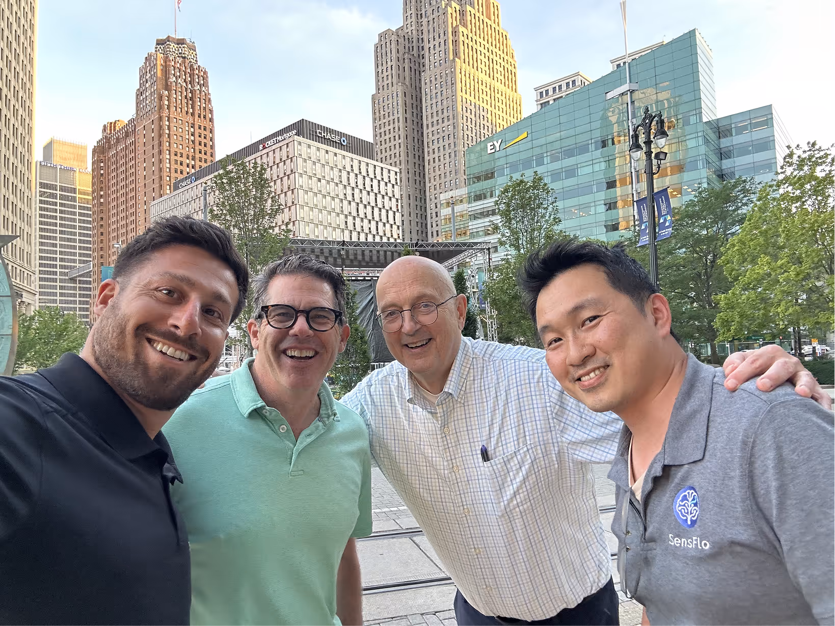 Four men smiling and posing for a selfie in an urban park with tall buildings in the background, including one with EY logo and another with Chase logo.