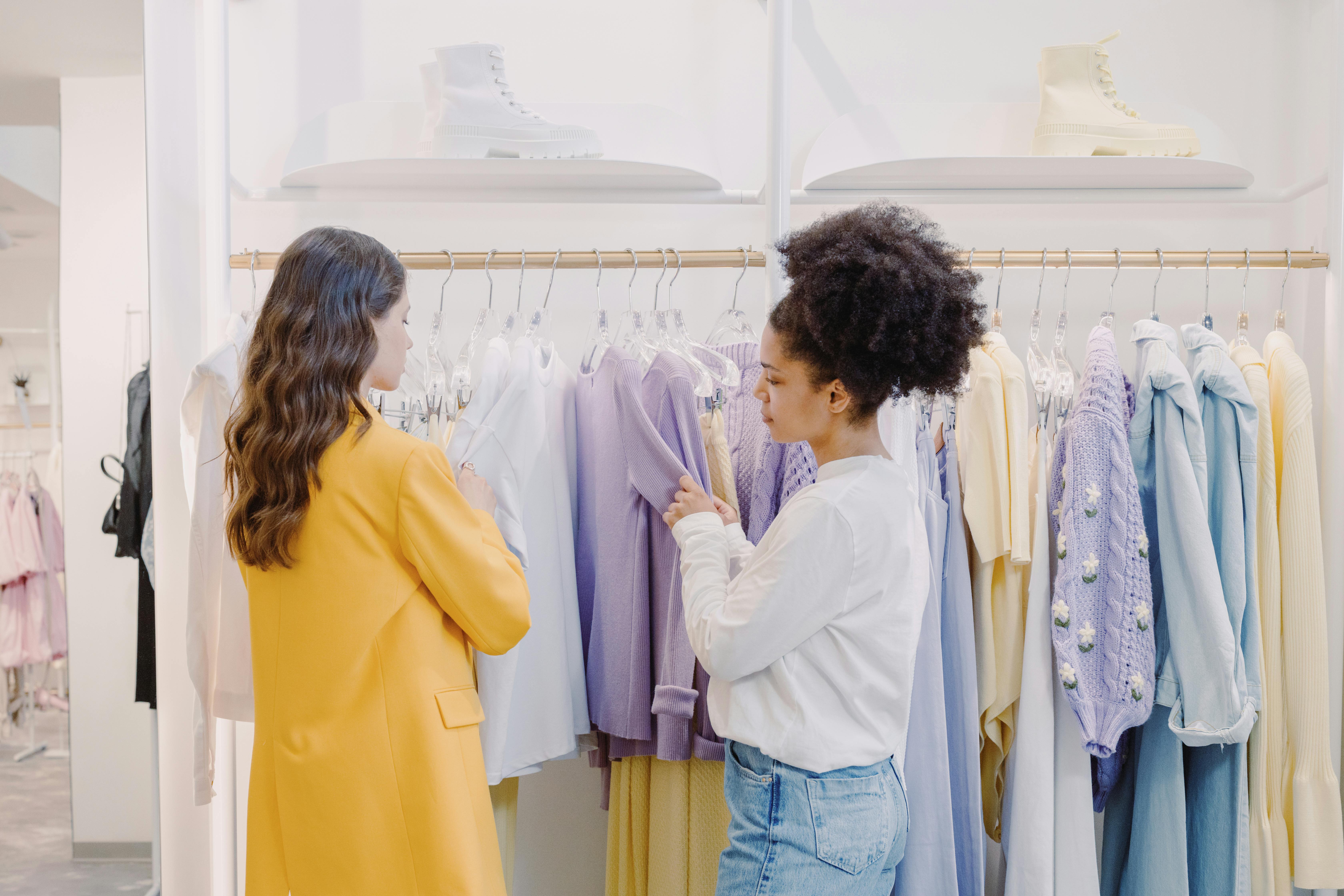 Fashion stylist consulting a client in a clothing store, selecting outfits from a wardrobe rack with pastel-colored dresses and jackets.