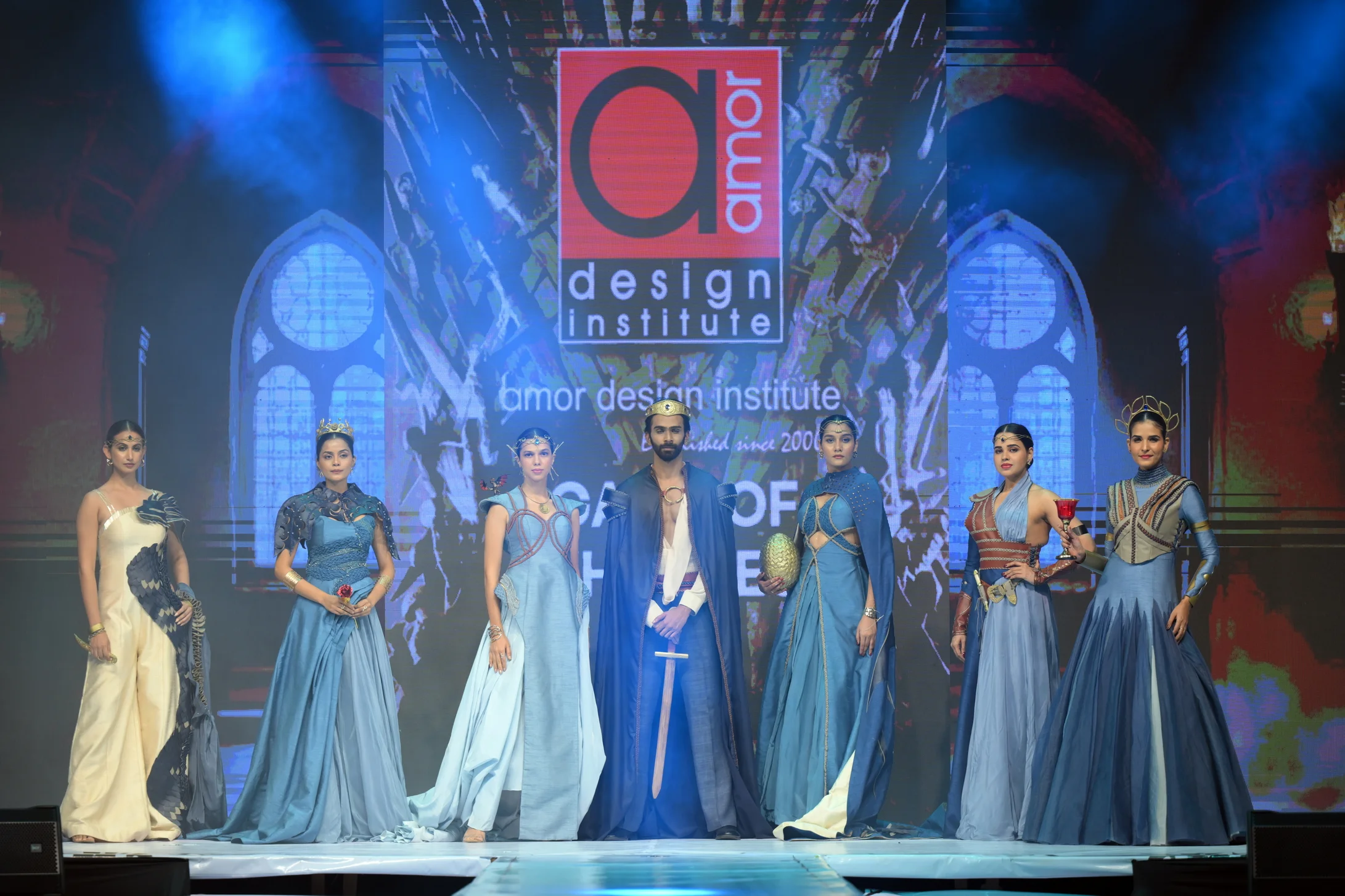 A group of models showcasing a fantasy-themed collection, including a central male model with a sword, on the runway at the Amor Glitzz fashion event hosted by Amor Design Institute.