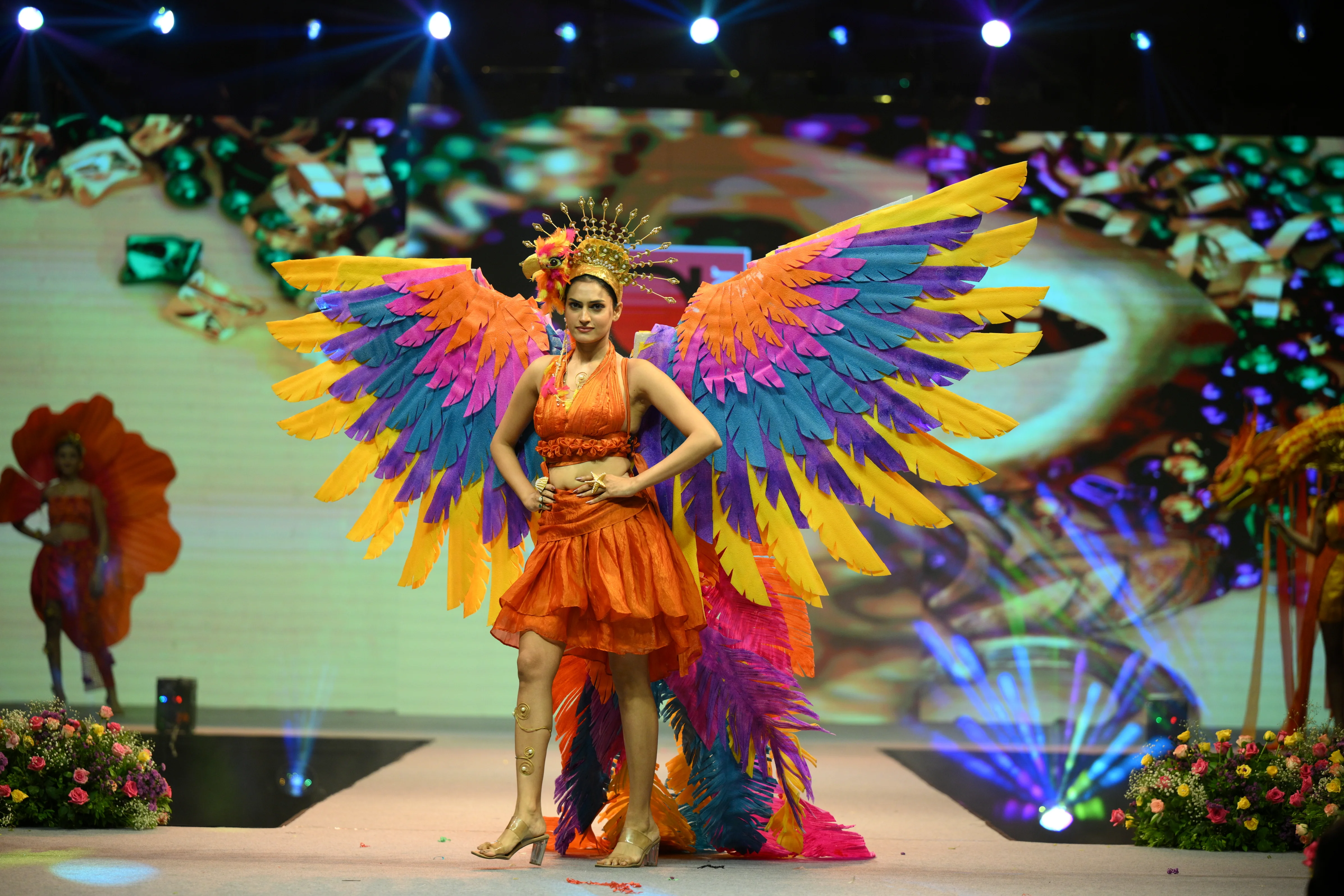 Fashion model walking the runway wearing a vibrant orange outfit with large, multi-colored feathered wings and a gold sunburst headpiece.