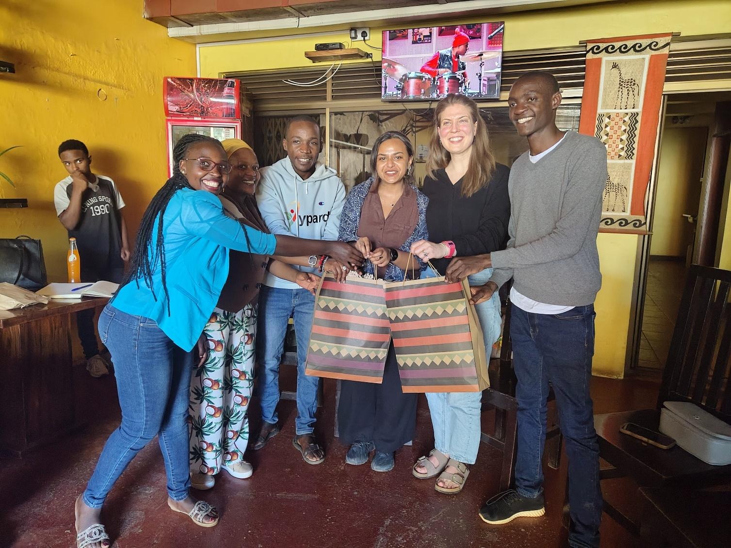 The image depicts a group of people gathered in a room with a colorful, patterned blanket on a table, with a backdrop featuring a mural and some decorations.AI-generated content may be incorrect.
