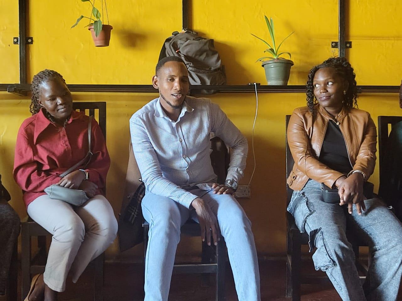 Three people are seated closely, facing each other, in a cozy, yellow-walled room with a potted plant on a table.AI-generated content may be incorrect.