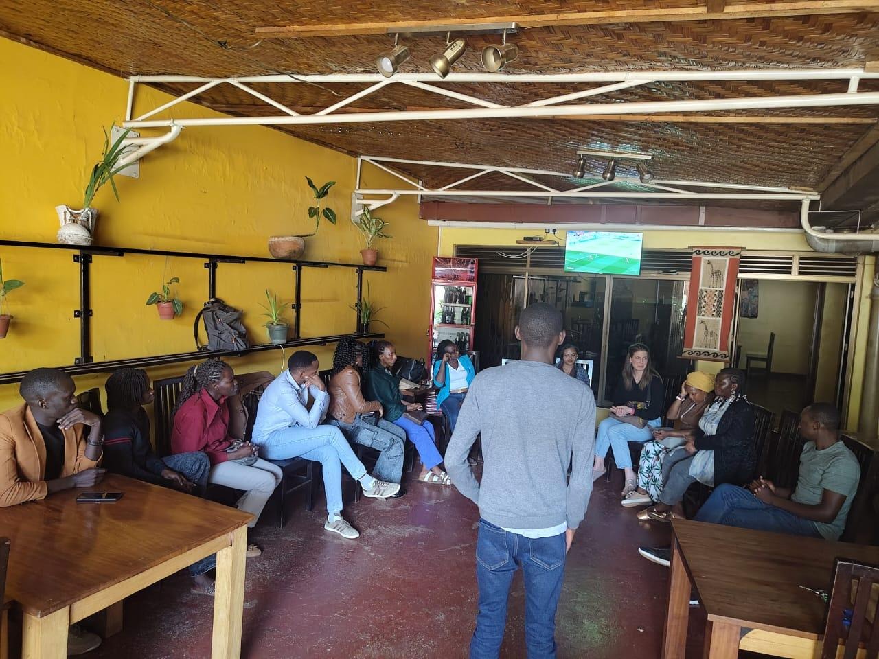 A group of people is gathered in a cozy, yellow-painted room with exposed beams, sitting at wooden tables, and some are engaged in conversation.AI-generated content may be incorrect.