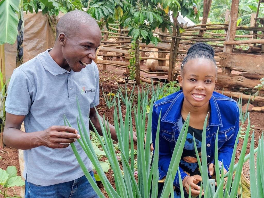 The image depicts two people, one in a dark blue shirt and the other in a blue plaid shirt, standing in a green, leafy garden, possibly engaged in gardening or planting activities.AI-generated content may be incorrect.