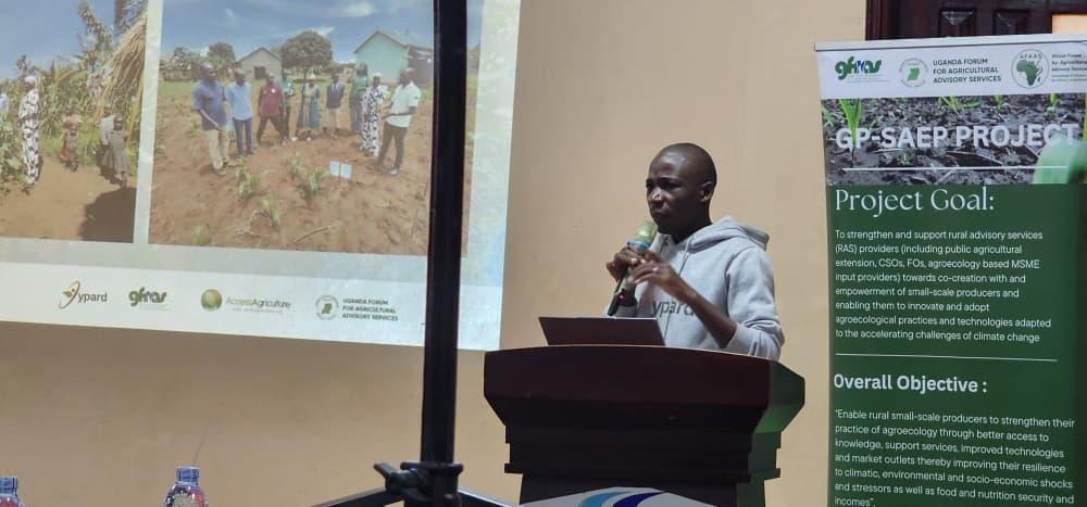 The image depicts a presentation at a forum in Uganda, focusing on the GP-SAEP Project's goal to enhance rural advisory services for small-scale producers, promoting agroecological practices and resilience against climate challenges.AI-generated content may be incorrect.